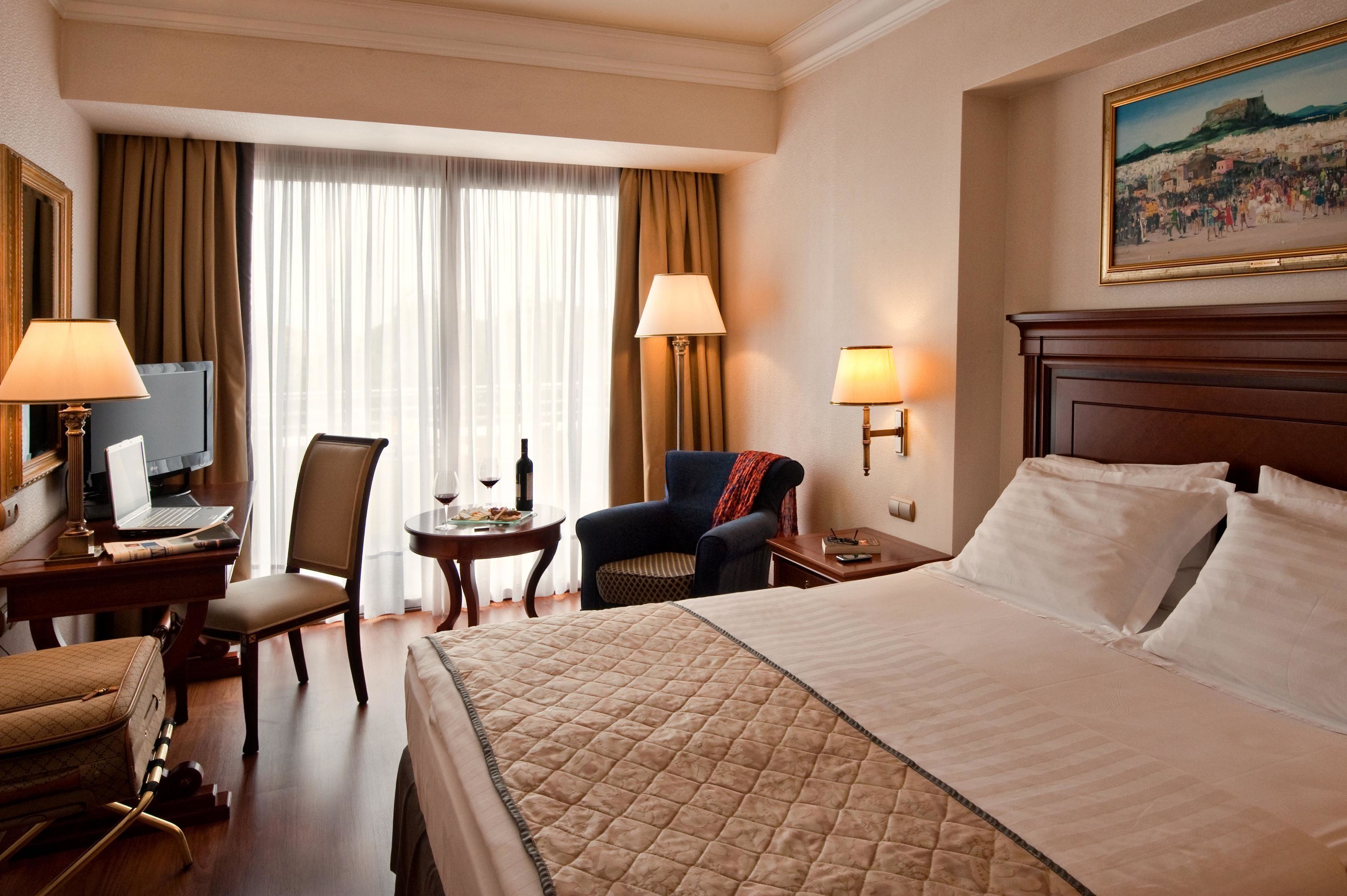 Une chambre d’hôtel avec un lit, deux chaises, un bureau avec un ordinateur portable, une table avec un verre de vin et un tableau au mur.