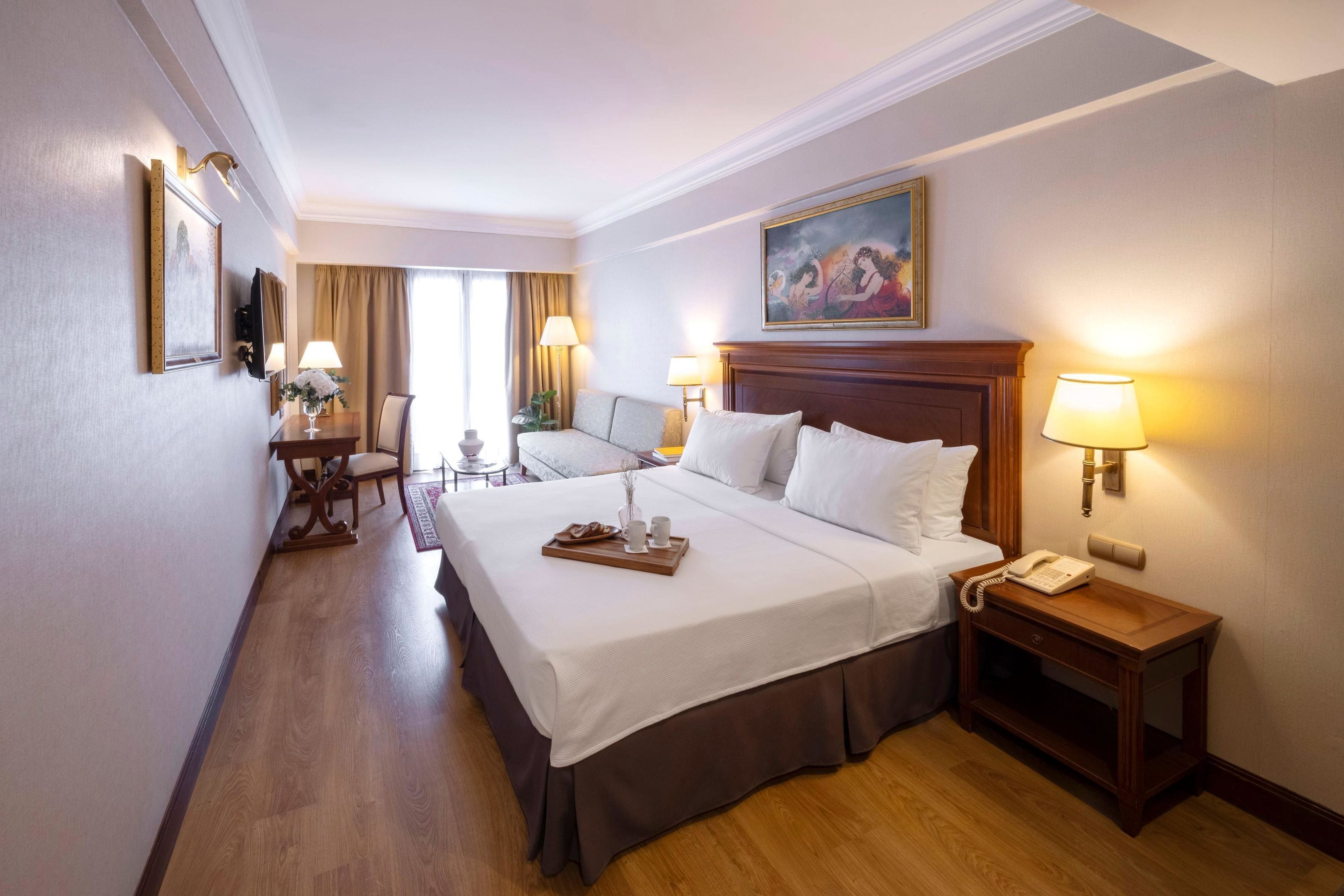 Habitación de hotel con una cama grande, mesitas de noche, un escritorio con una silla, un cuadro en la pared y una ventana con cortinas.