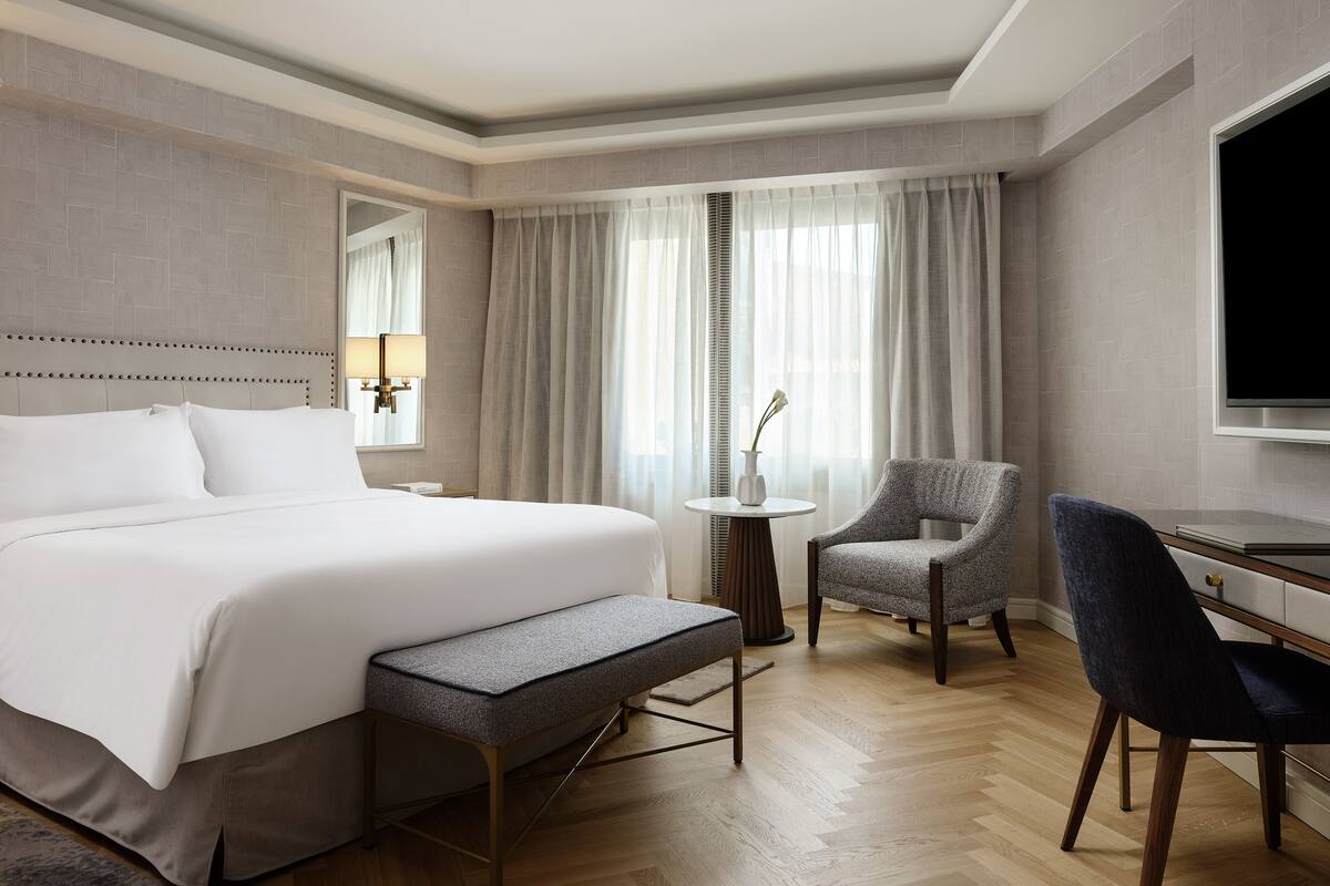 A hotel room with a bed, a desk, two chairs, a small table with a vase, and a television.