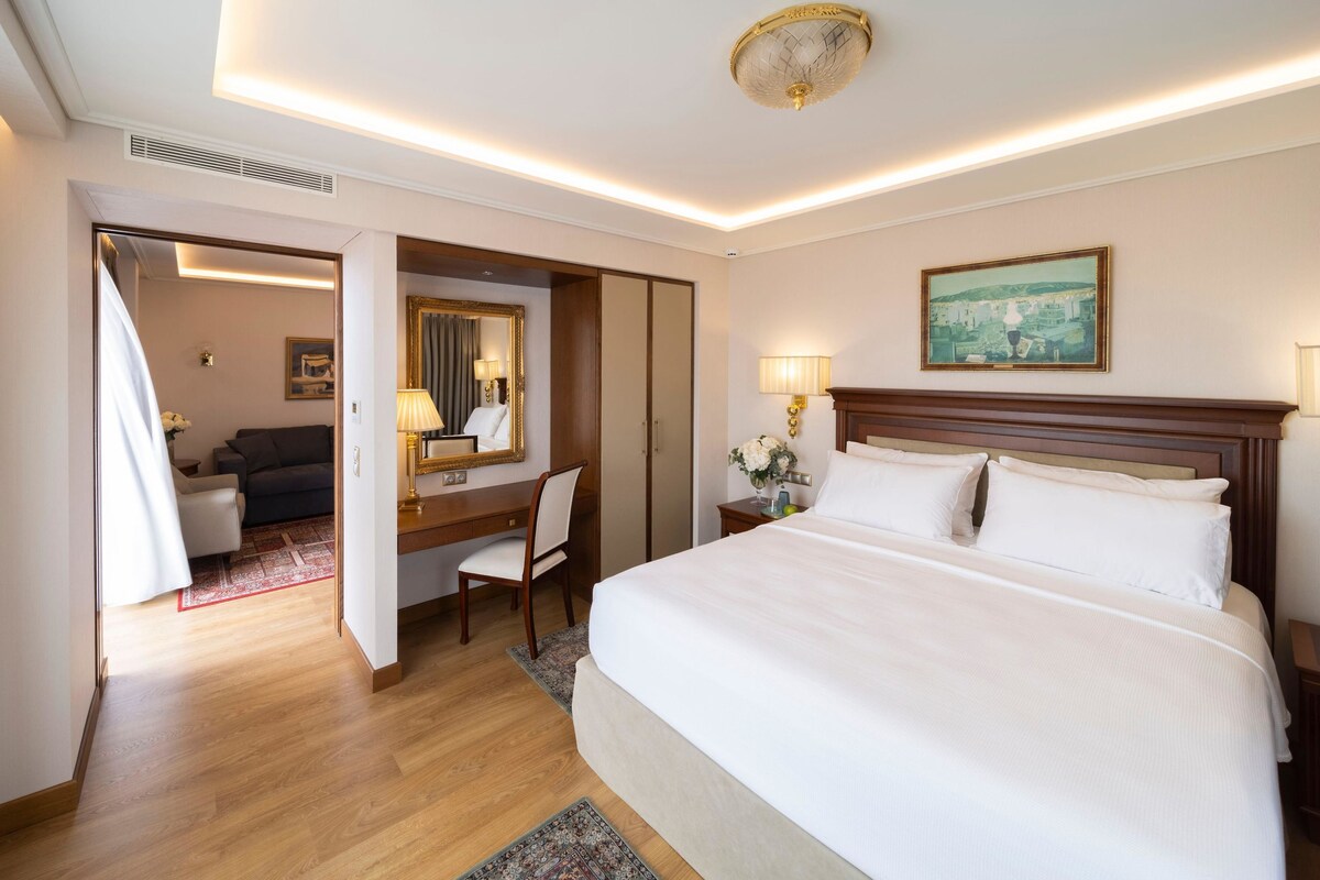 A hotel room with a large bed, a desk with a chair, a mirror, a painting on the wall, and a view of the cityscape.