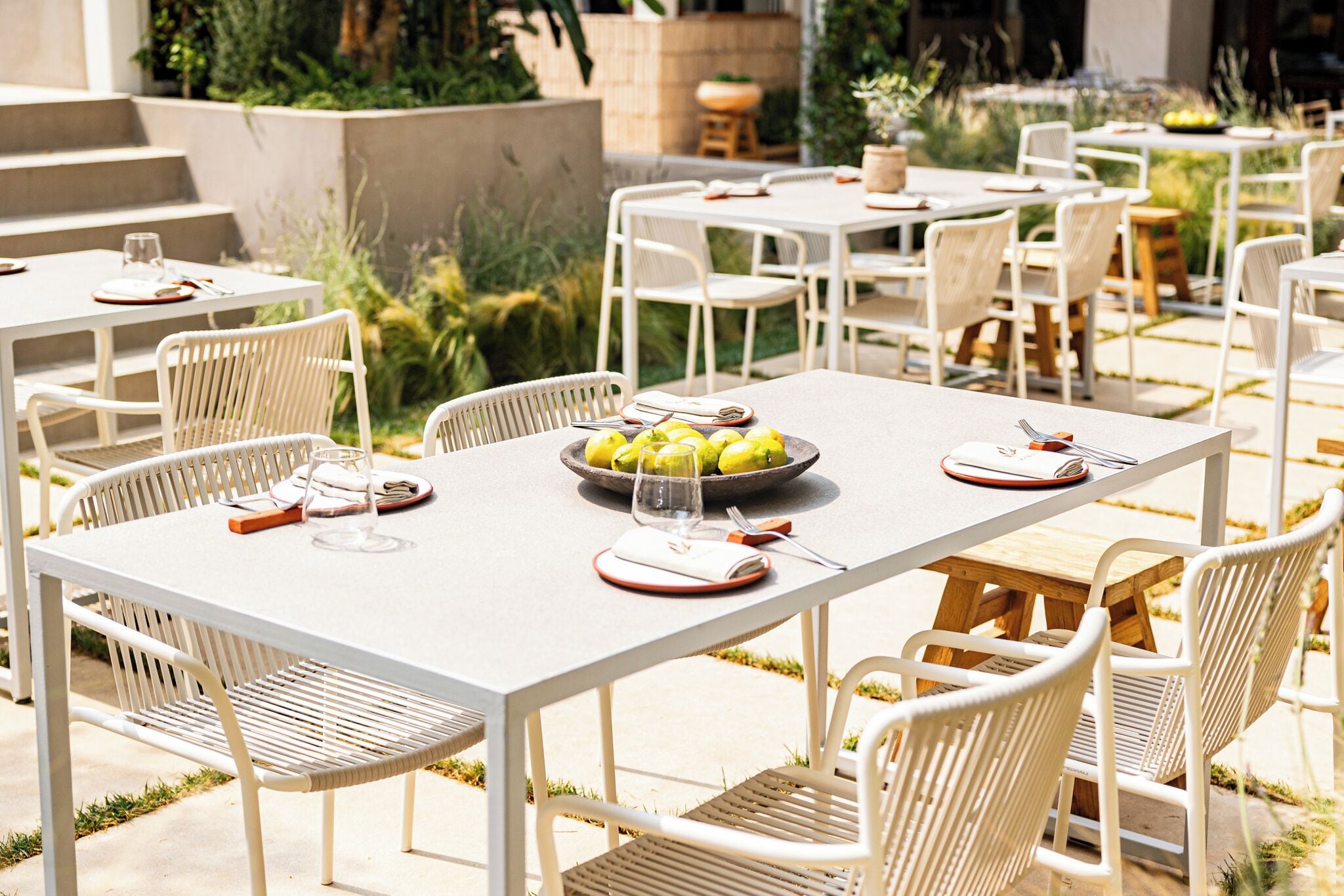 An outdoor dining area with white metal tables and chairs, a bowl of lemons, and a wine glass on one table.
