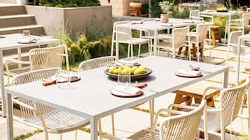 An outdoor dining area with white metal tables and chairs, a bowl of lemons, and a wine glass on one table.