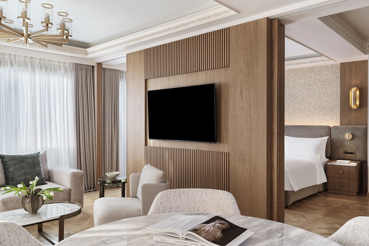 A hotel room with a flat-screen TV mounted on a wooden panel wall, a bed with a headboard, a seating area with a round table, and a large window with curtains.
