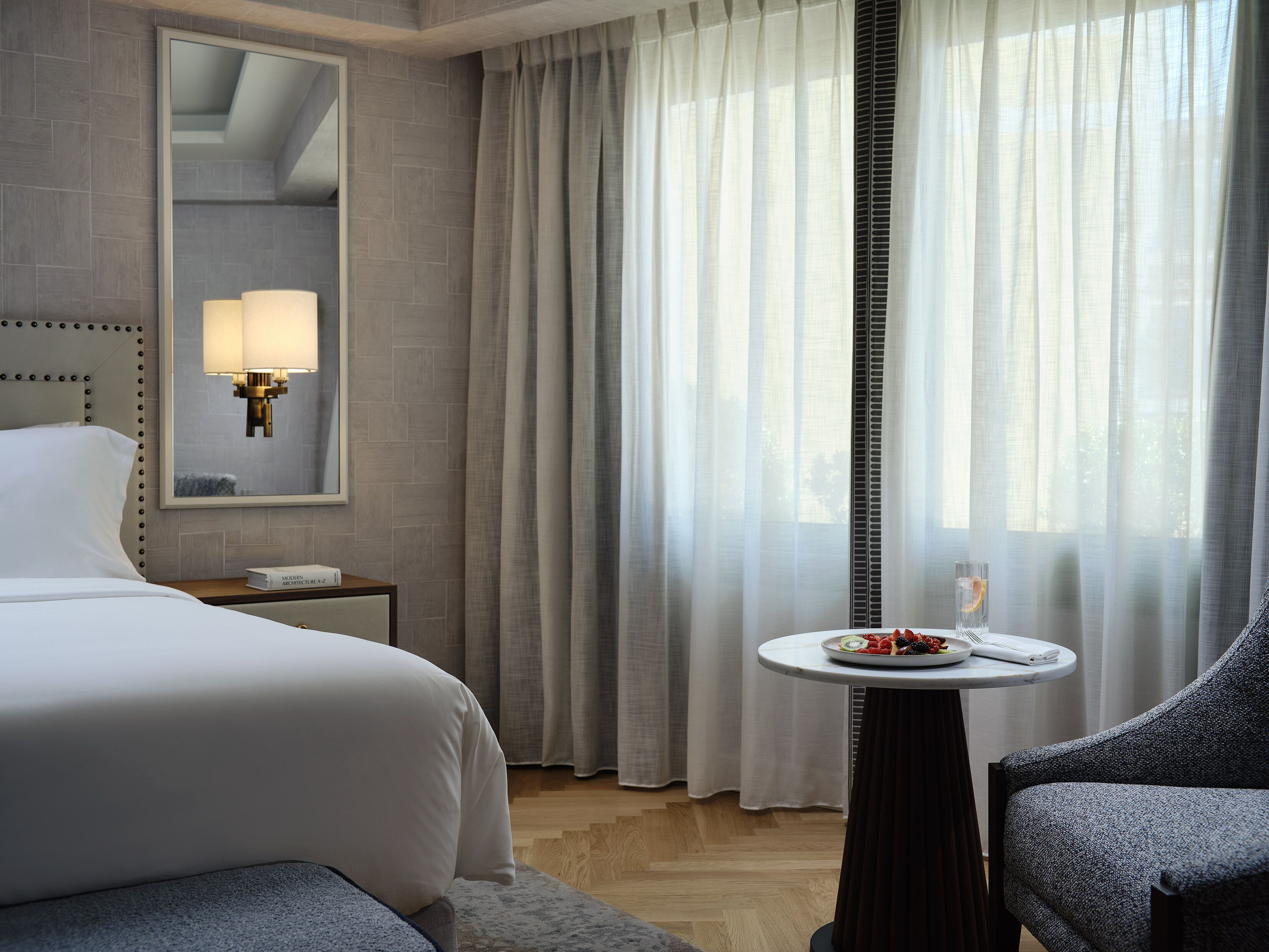 Ein Hotelzimmer mit einem Bett, einem Nachttisch mit Lampe, einem kleinen runden Tisch mit Obstteller und einem Sessel.