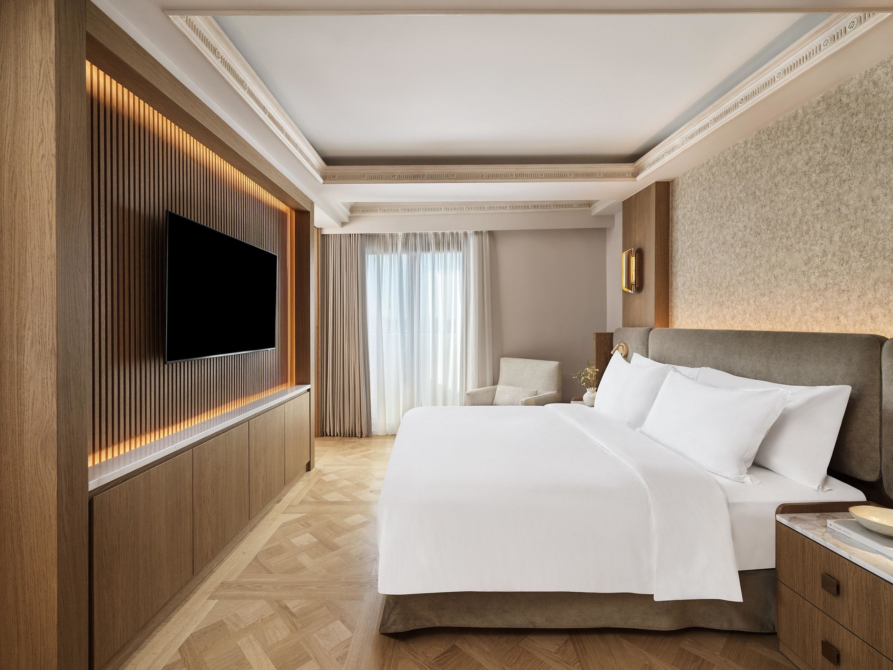 A hotel room with a large bed, a flat-screen TV mounted on the wall, a wooden panel design, and a large window with curtains.