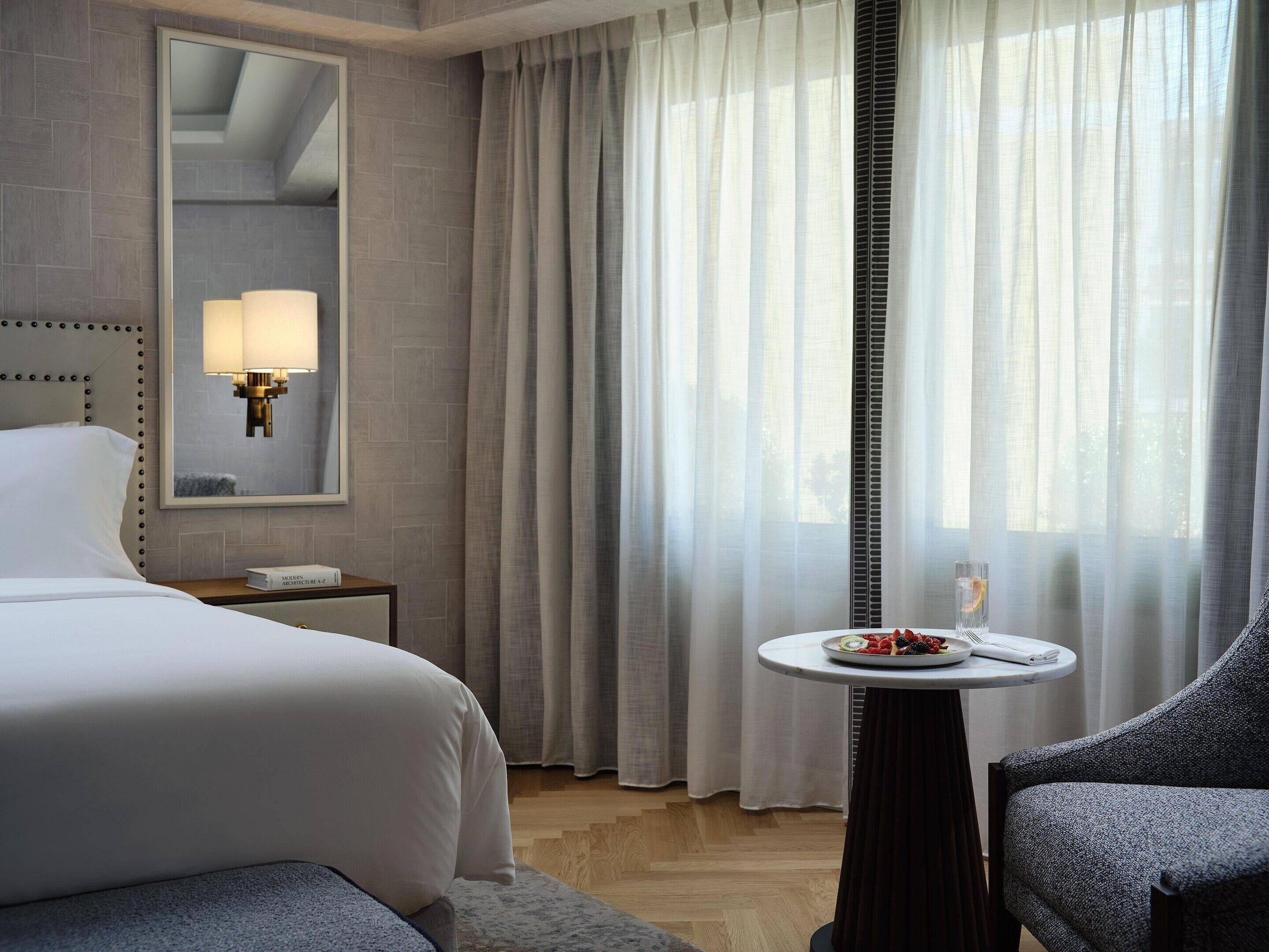 Une chambre d’hôtel avec un lit, une table de chevet avec une lampe, une petite table ronde sur laquelle se trouve une assiette de fruits, et une chaise.