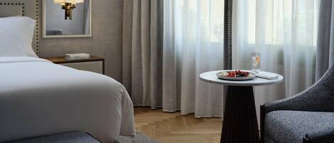 A hotel room with a bed, a bedside table with a lamp, a small round table with a plate of fruit, and a chair.