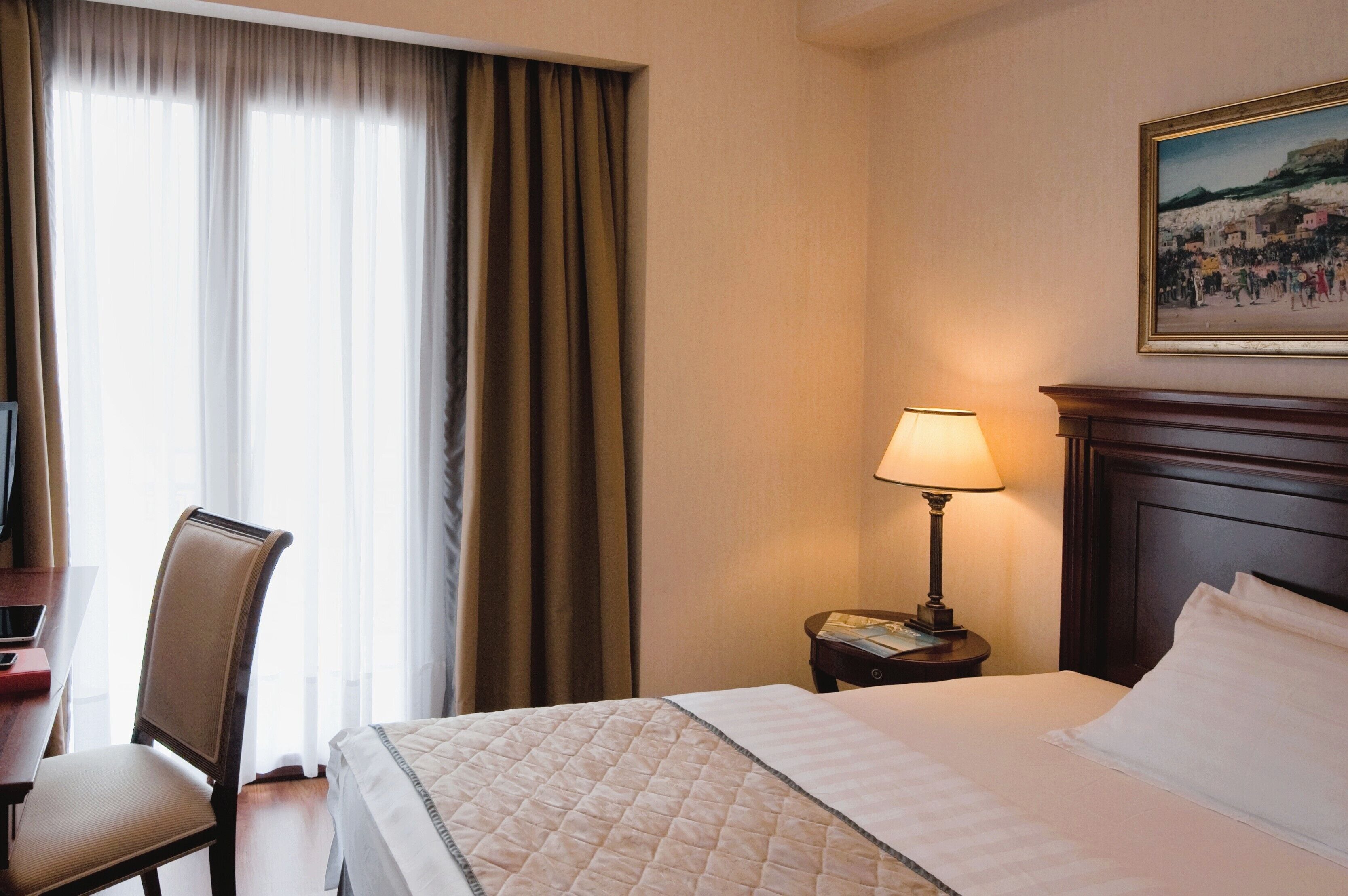 A hotel room with a bed, a desk, a chair, a lamp, and a painting on the wall.