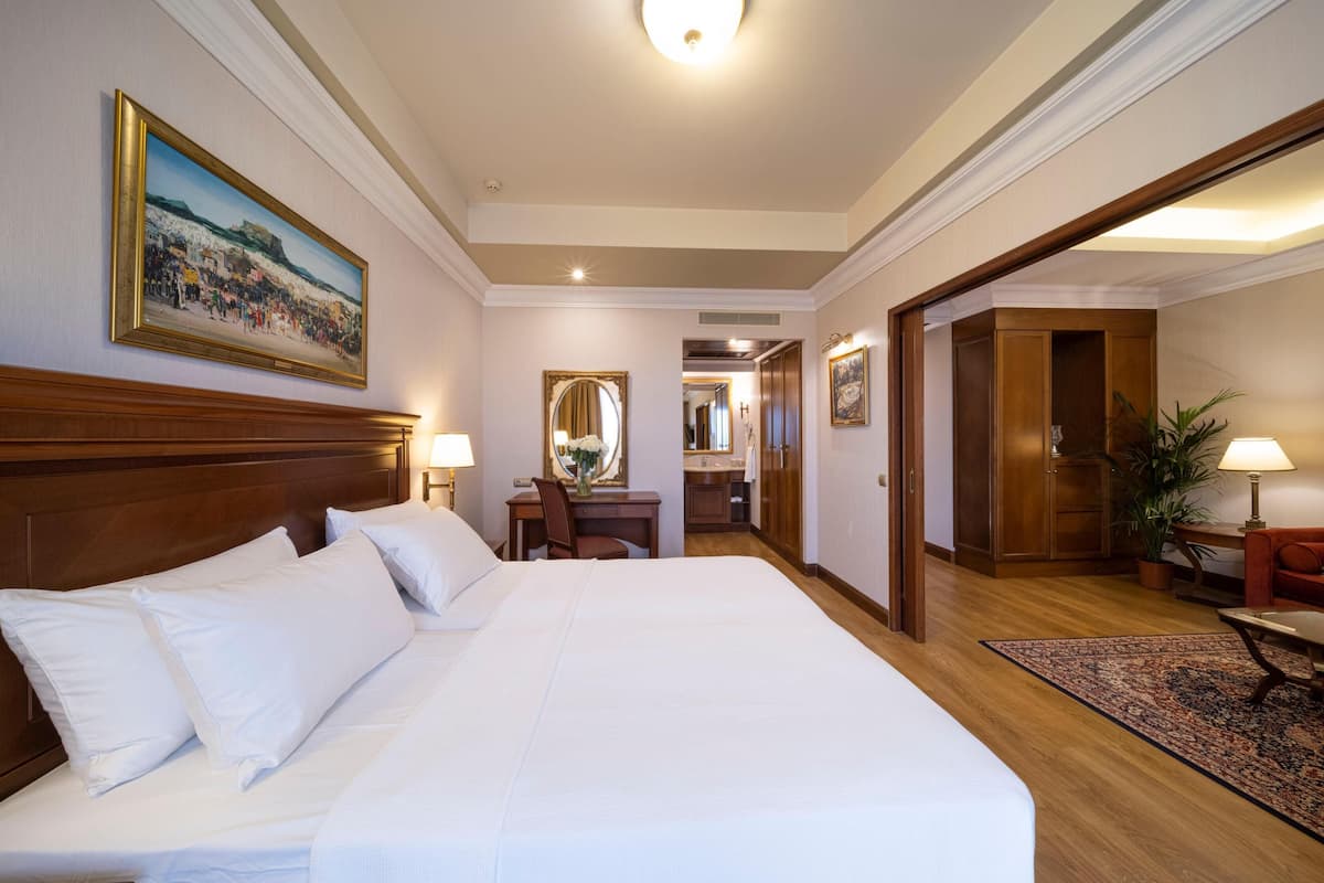 A hotel room with a large bed, a desk with a chair, a mirror, a bathroom, and a dining area.
