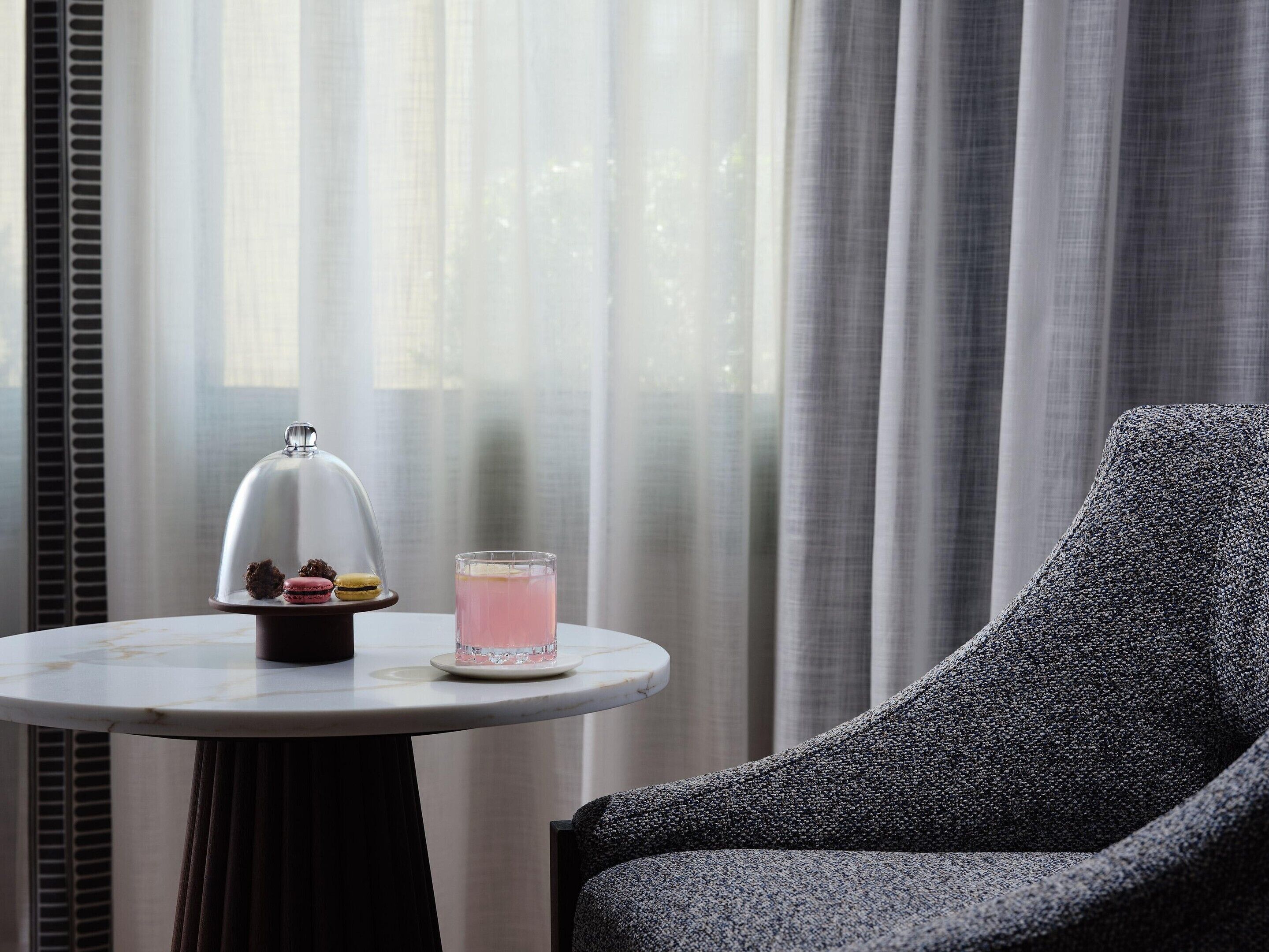 A round table with a glass of pink drink and a dessert display with macarons and a chocolate truffle. A grey upholstered armchair is positioned to the right.