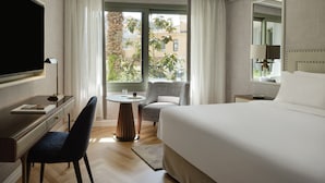 Habitación de hotel con cama, escritorio con silla, televisión, ventana con cortinas y vista a edificios.