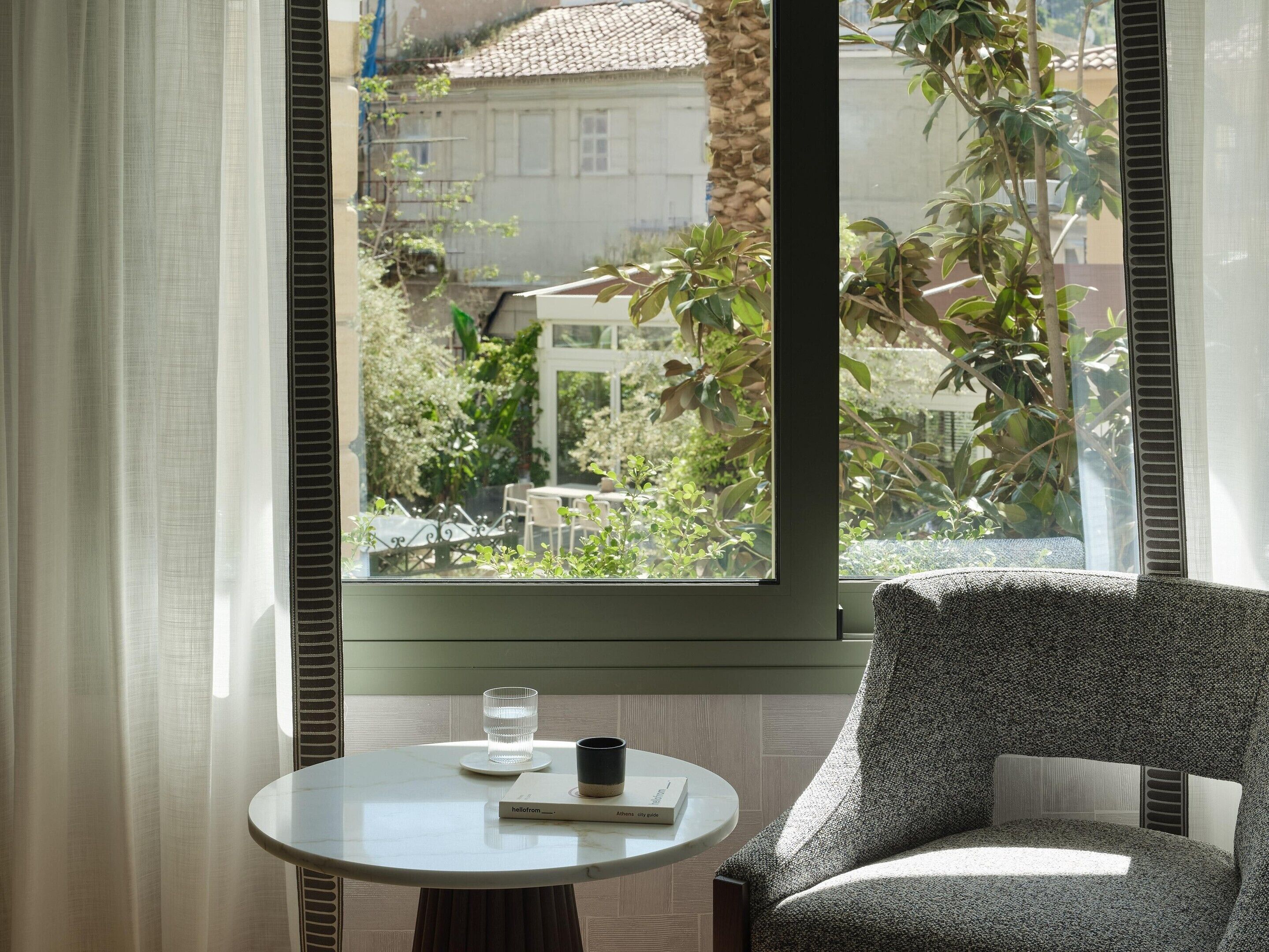 Um quarto com uma mesa redonda, uma cadeira, uma janela com vista para vegetação e edifícios, e um livro intitulado 'hello from Athens city guide'.