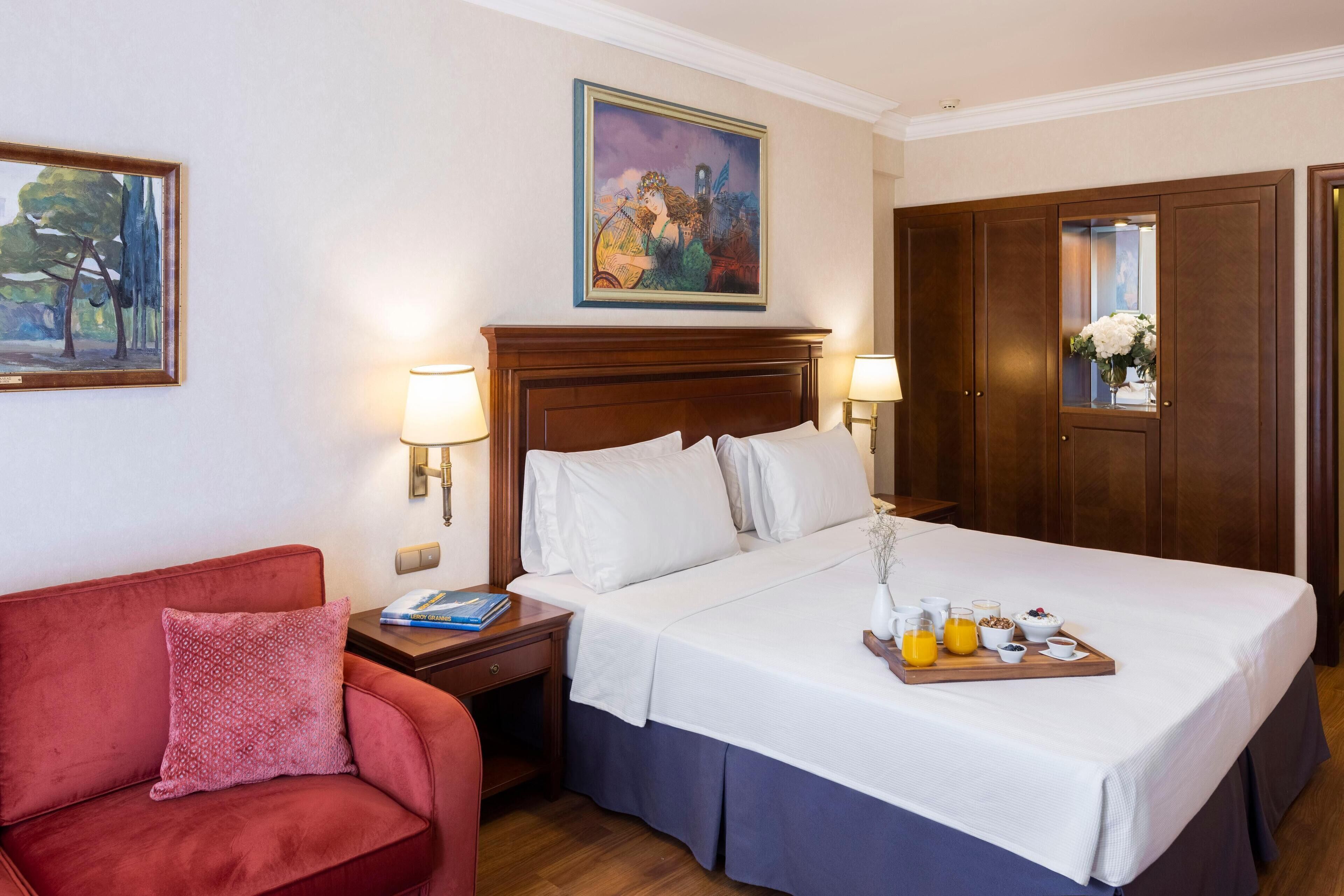 Habitación de hotel con una cama grande, un sofá rojo, una mesita de noche con lámpara, un cuadro en la pared y un armario de madera.
