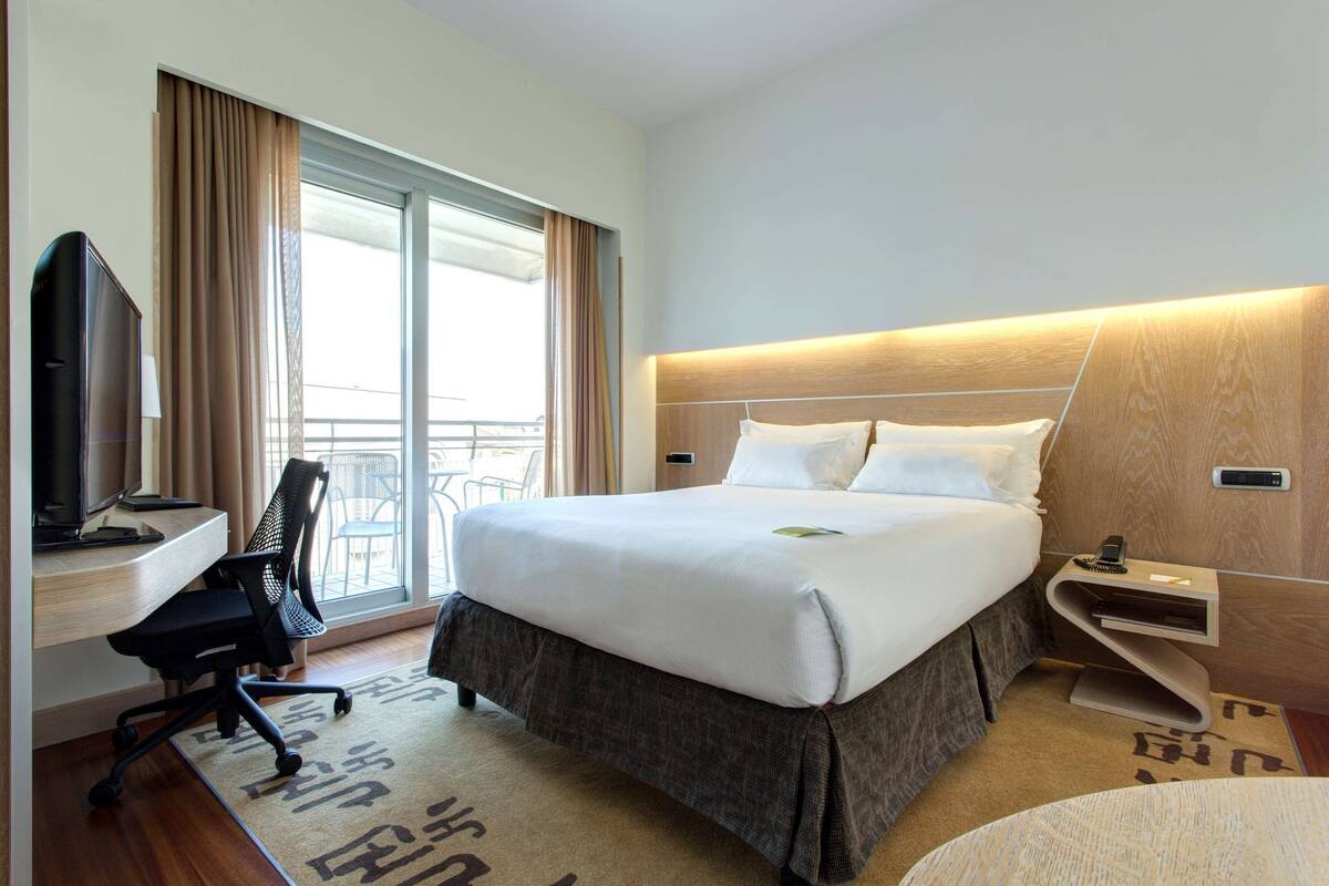 A hotel room with a large bed, a desk with a chair, a television, and a balcony with curtains.
