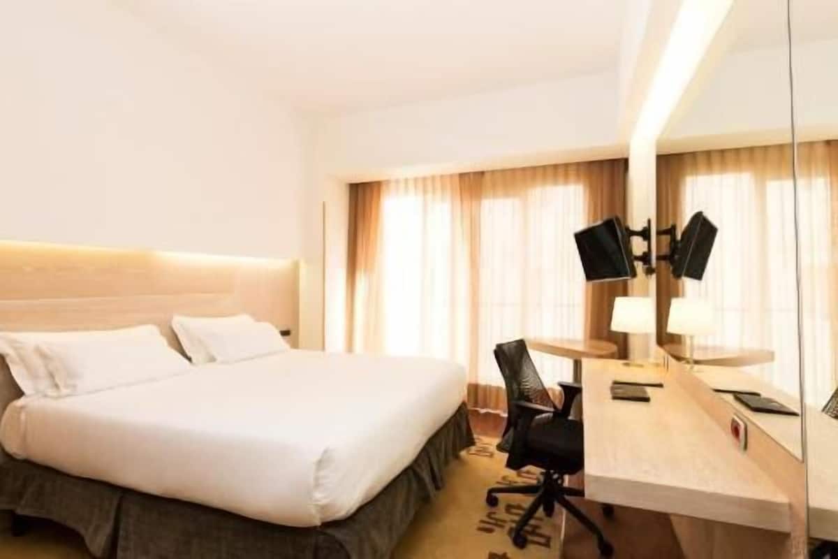 A hotel room with a bed, desk, chair, and a large mirror.