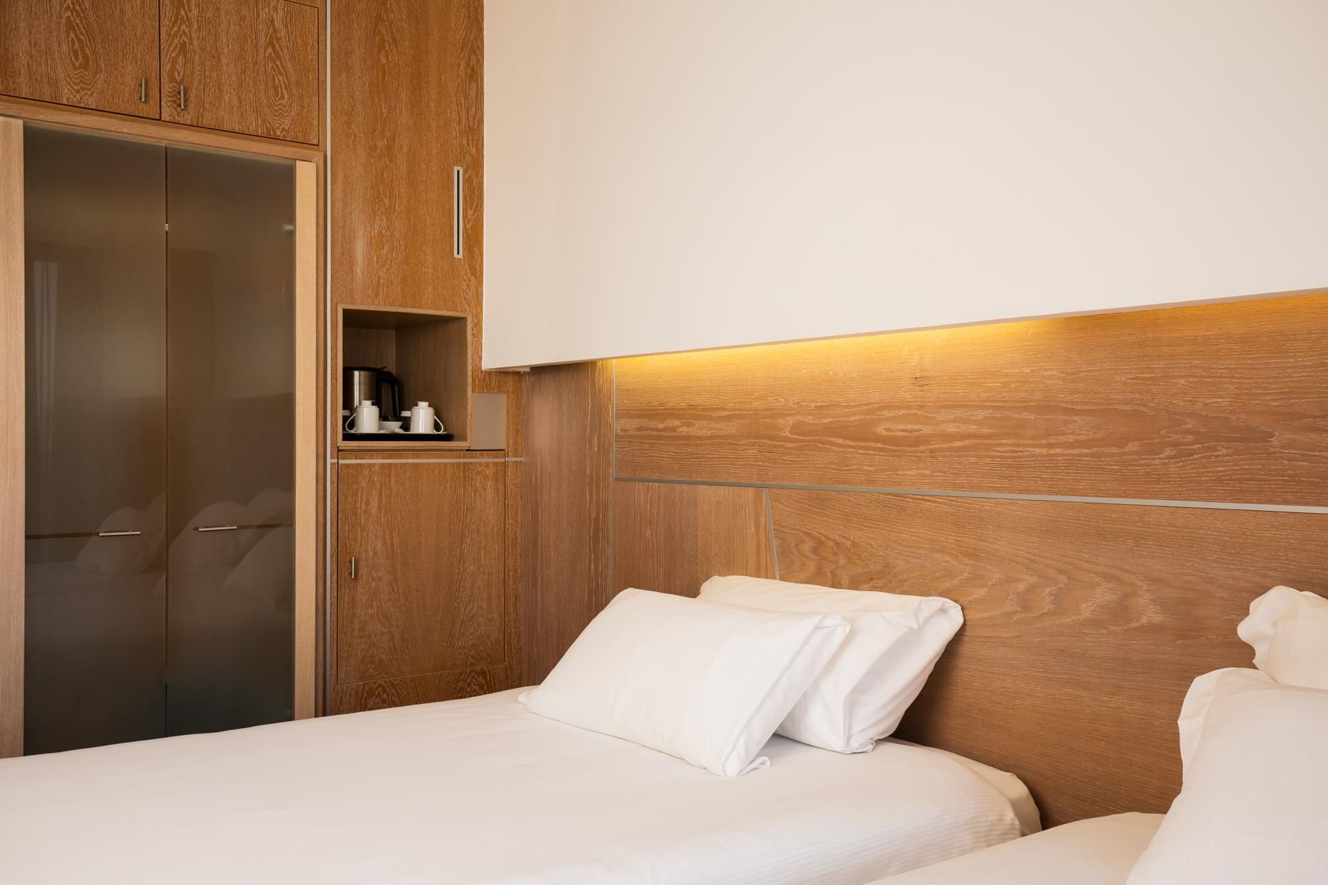 Habitación de hotel con cabecera de madera, armario con espejo y mesita de noche con tetera y tazas.