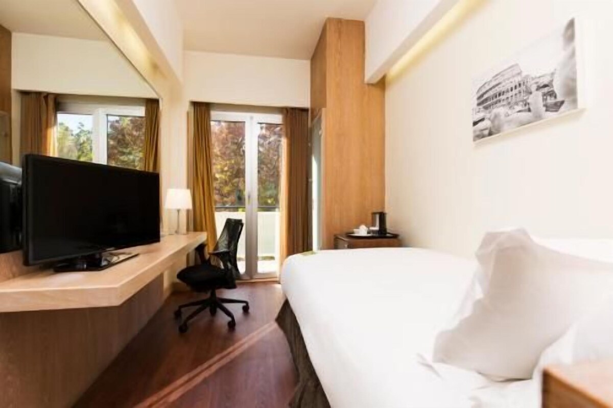 A hotel room with a bed, a desk with a television, a chair, and a window with a view of trees.