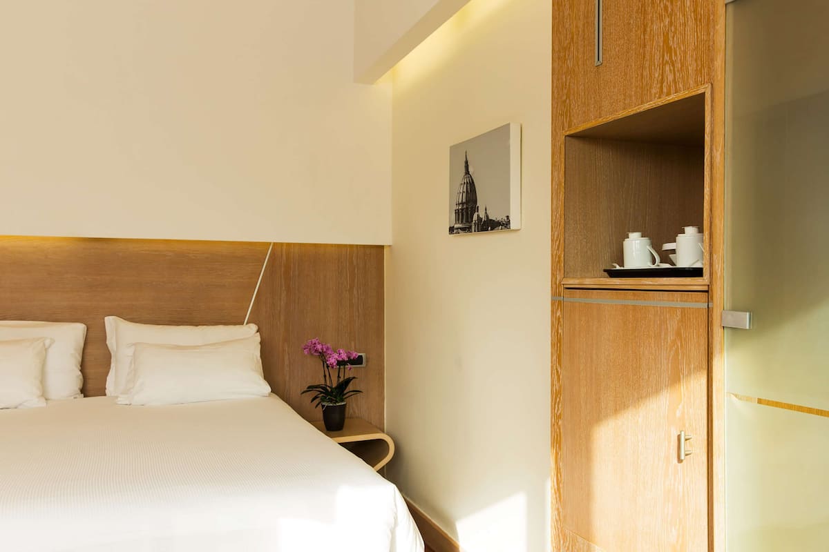 A hotel room with a bed, a wooden headboard, a small table with a potted plant, and a cabinet with kitchen items.