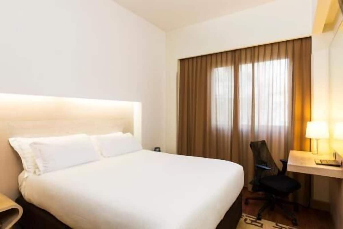 A hotel room with a bed, a desk, a chair, and a window with curtains.