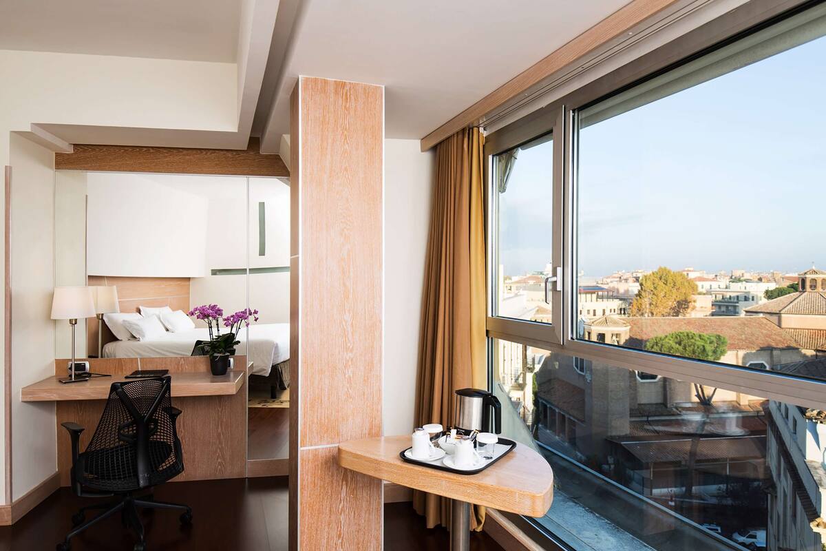 A hotel room with a bed, bedside table, desk, chair, and a large window offering a city view.