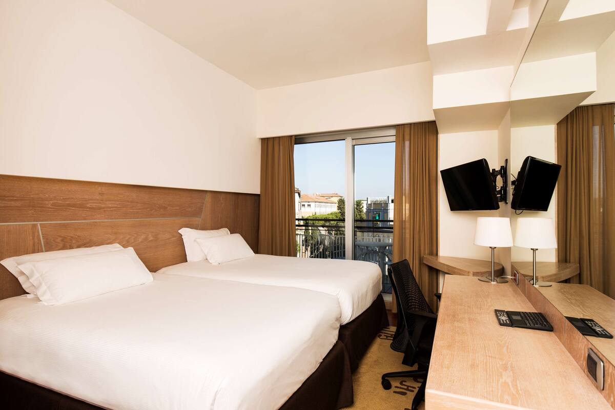A hotel room with two beds, a desk with a computer, a chair, a TV, and a balcony with a view of buildings.