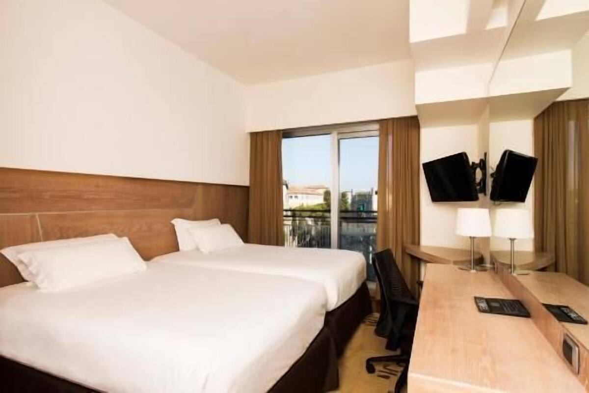 A hotel room with two beds, a desk, a chair, a TV, and a view of the city.