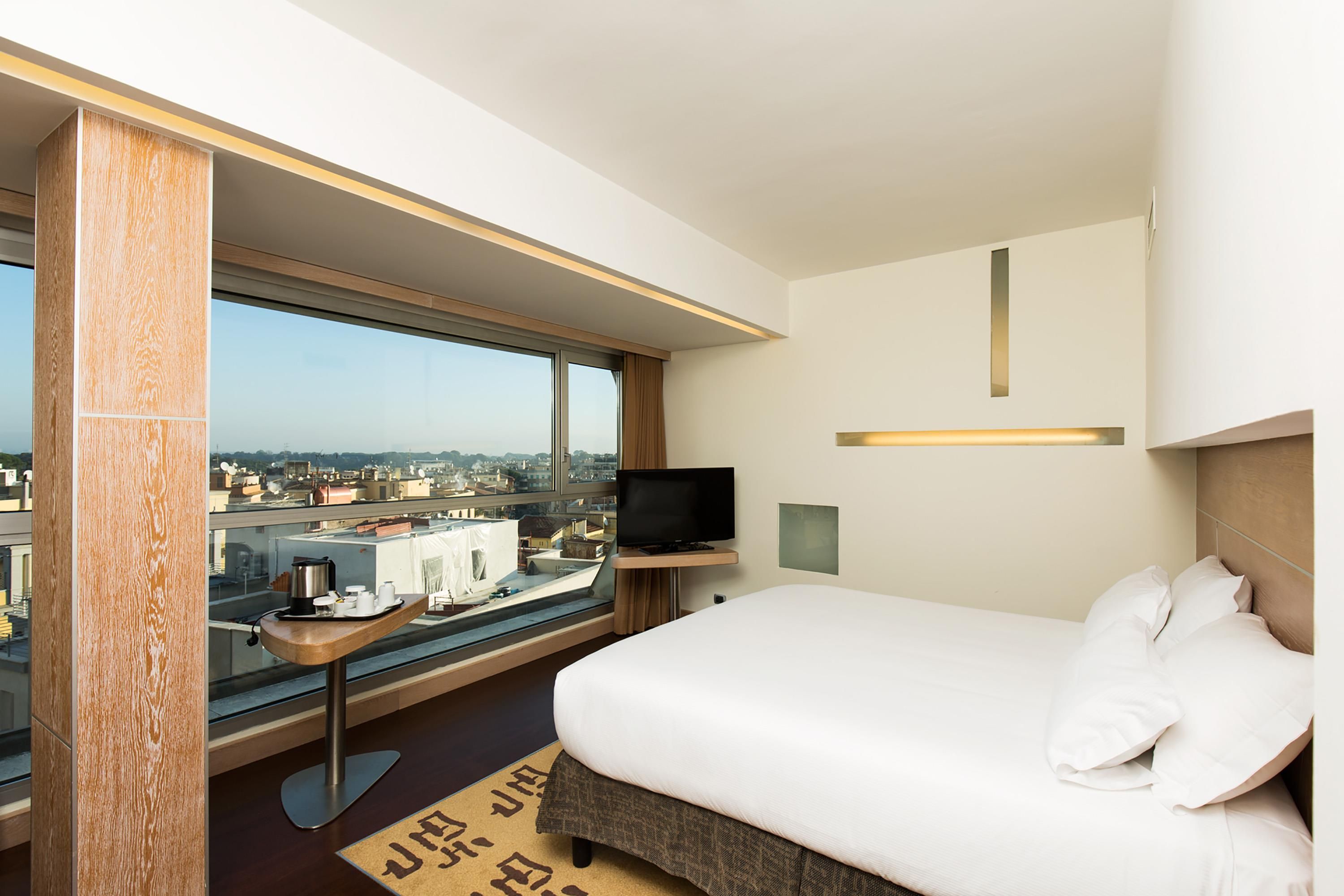 A hotel room with a large window, a bed, a TV, and a small table with a tray of items.