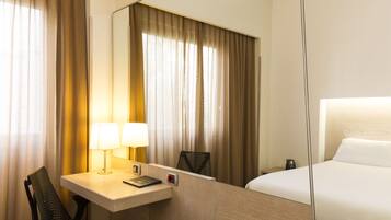 A hotel room with a bed, a desk with a lamp, a chair, and a large mirror.