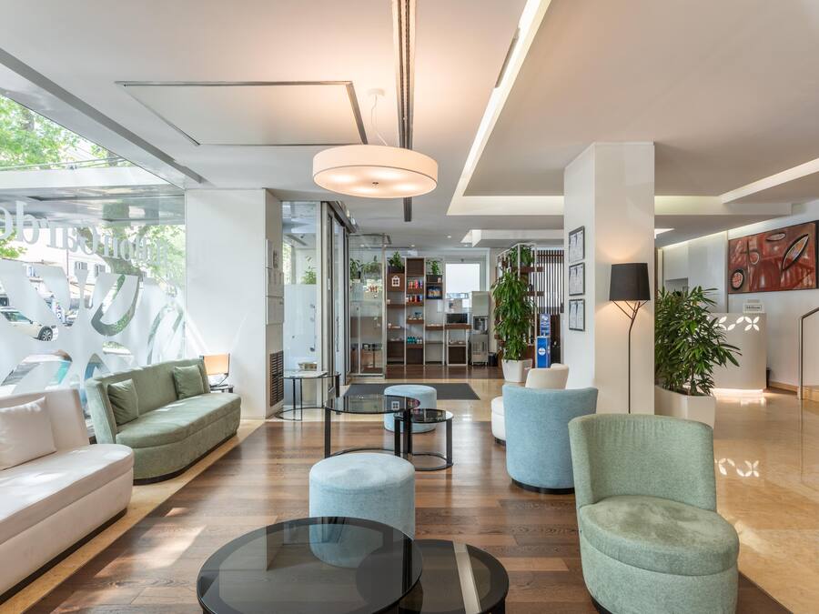 Un moderno vestíbulo de hotel con un sofá, reposapiés y sillas, una mesa de centro de vidrio y un mostrador de recepción.
