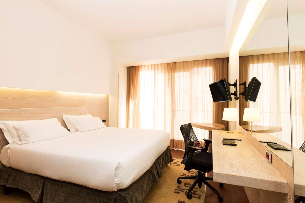 A hotel room with a large bed, a desk with a chair, a flat-screen TV, and a mirror.
