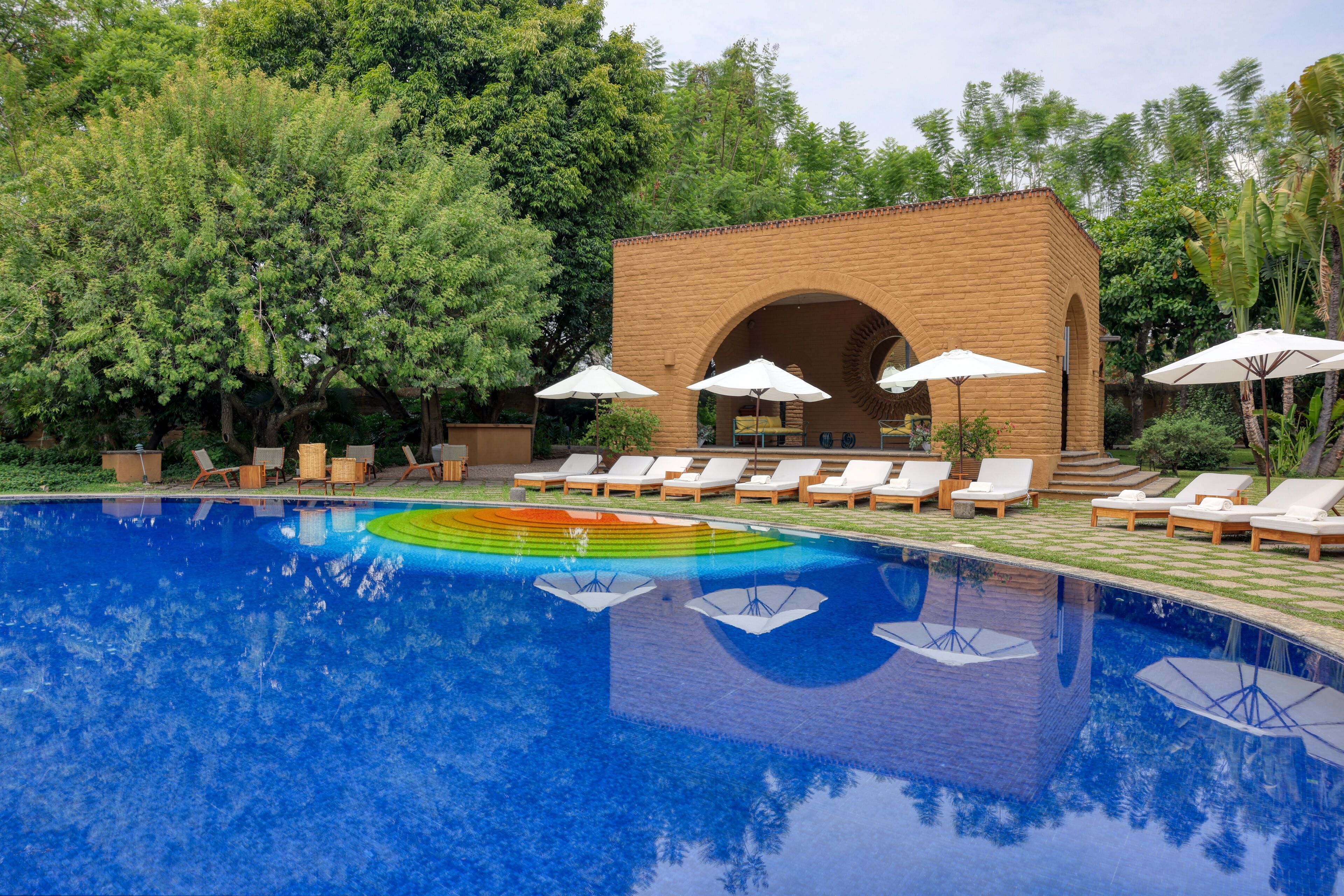 A swimming pool with lounge chairs and umbrellas, surrounded by trees and a brick building.