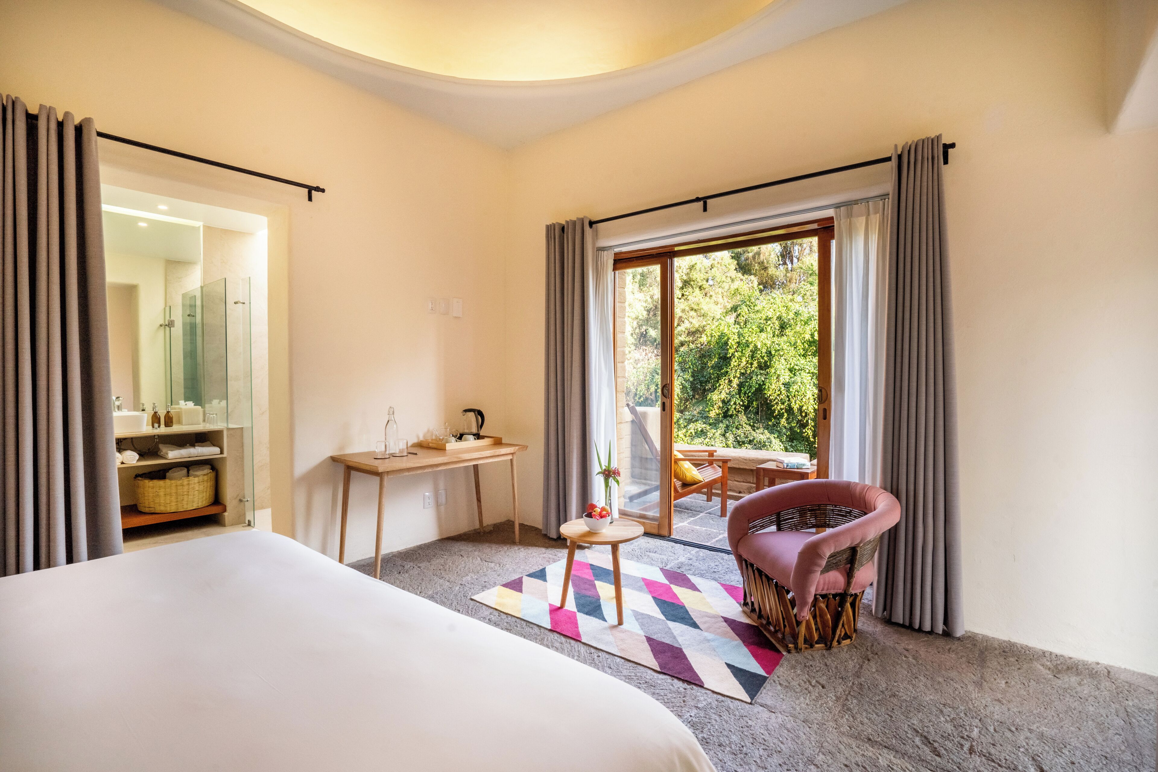 A hotel room with a bed, a small table with a vase, a chair, and a view of the outdoors.