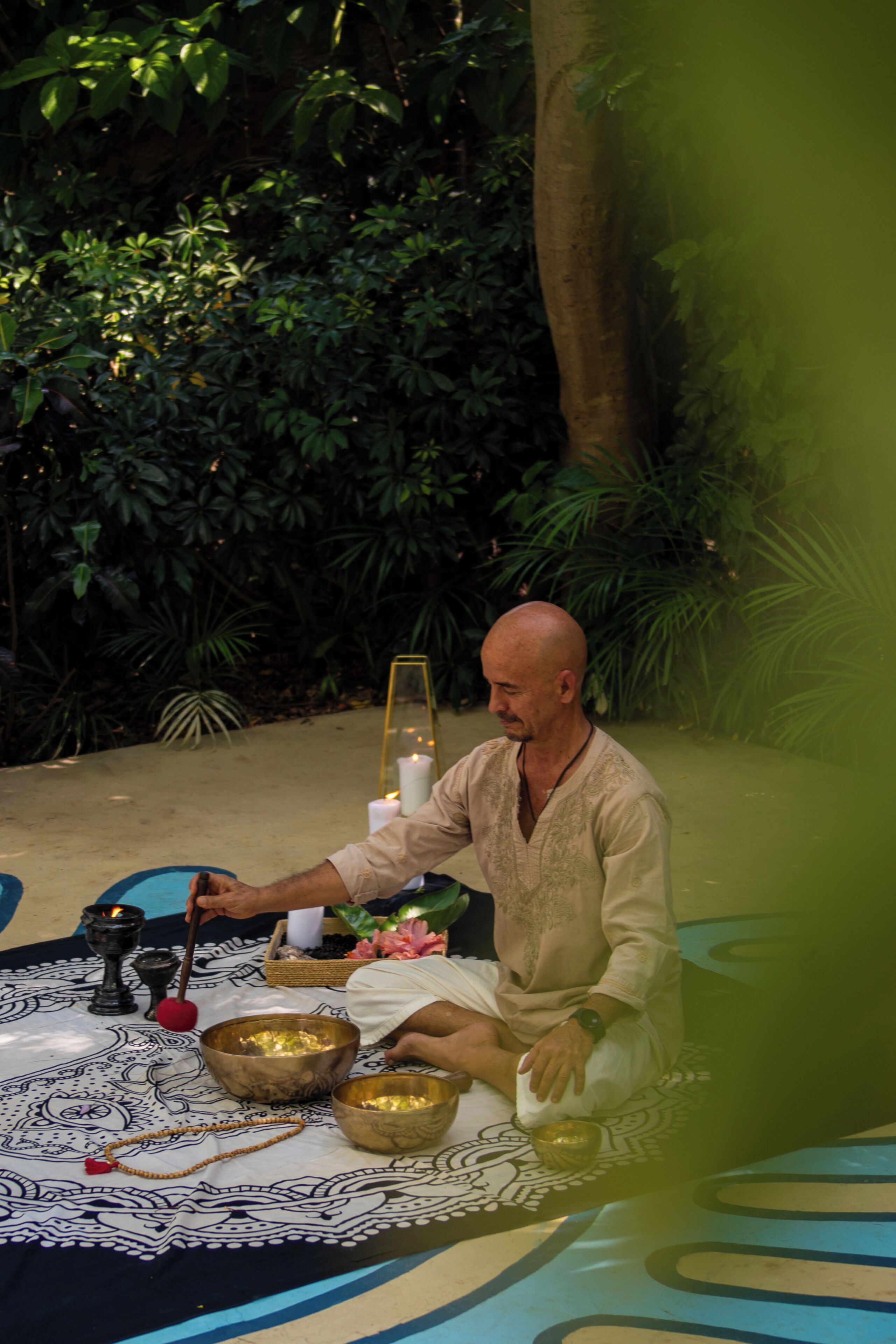 A person sitting on a mat with ritual items, surrounded by greenery.