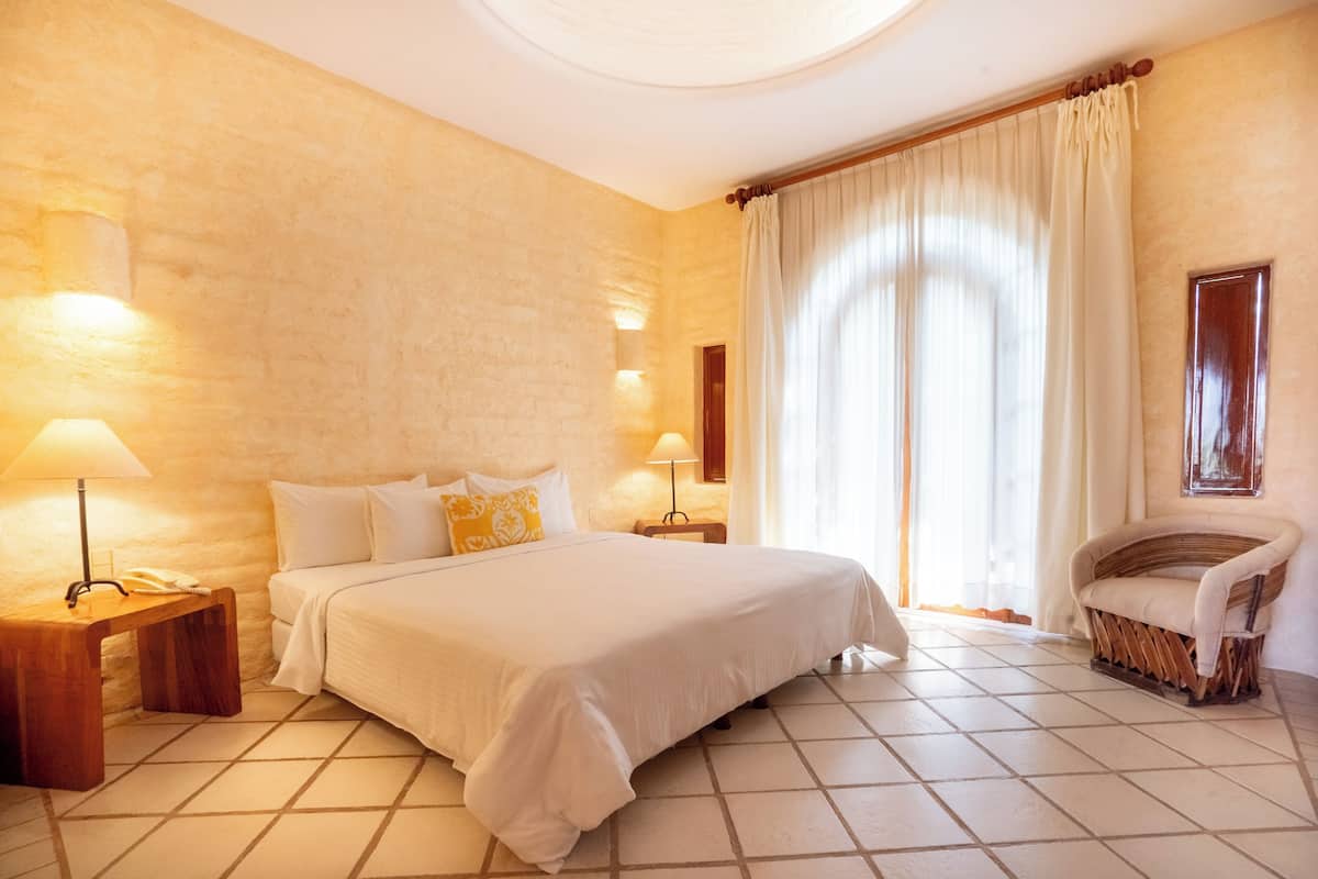 A hotel room with a large bed, bedside lamps, a wicker chair, and a view of the outdoors.