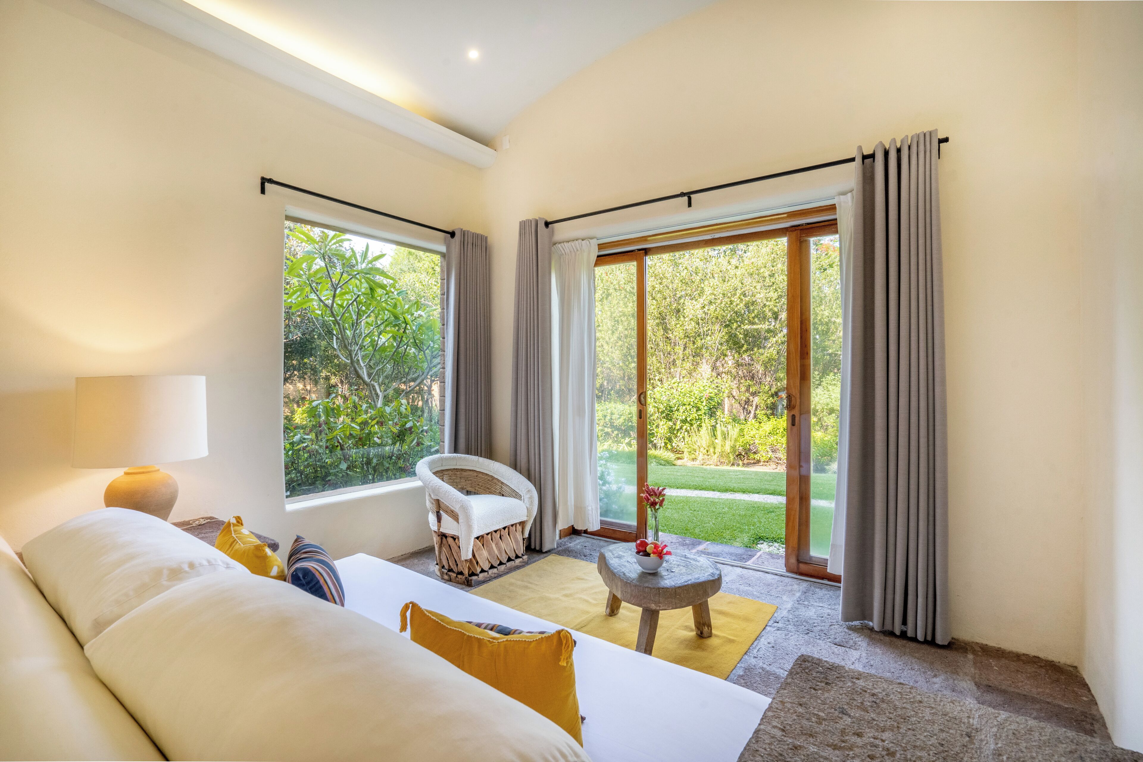 A room with a bed, a chair, a table, and a view of greenery through large windows.