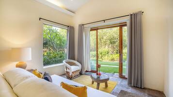 A room with a bed, a chair, a table, and a view of greenery through large windows.