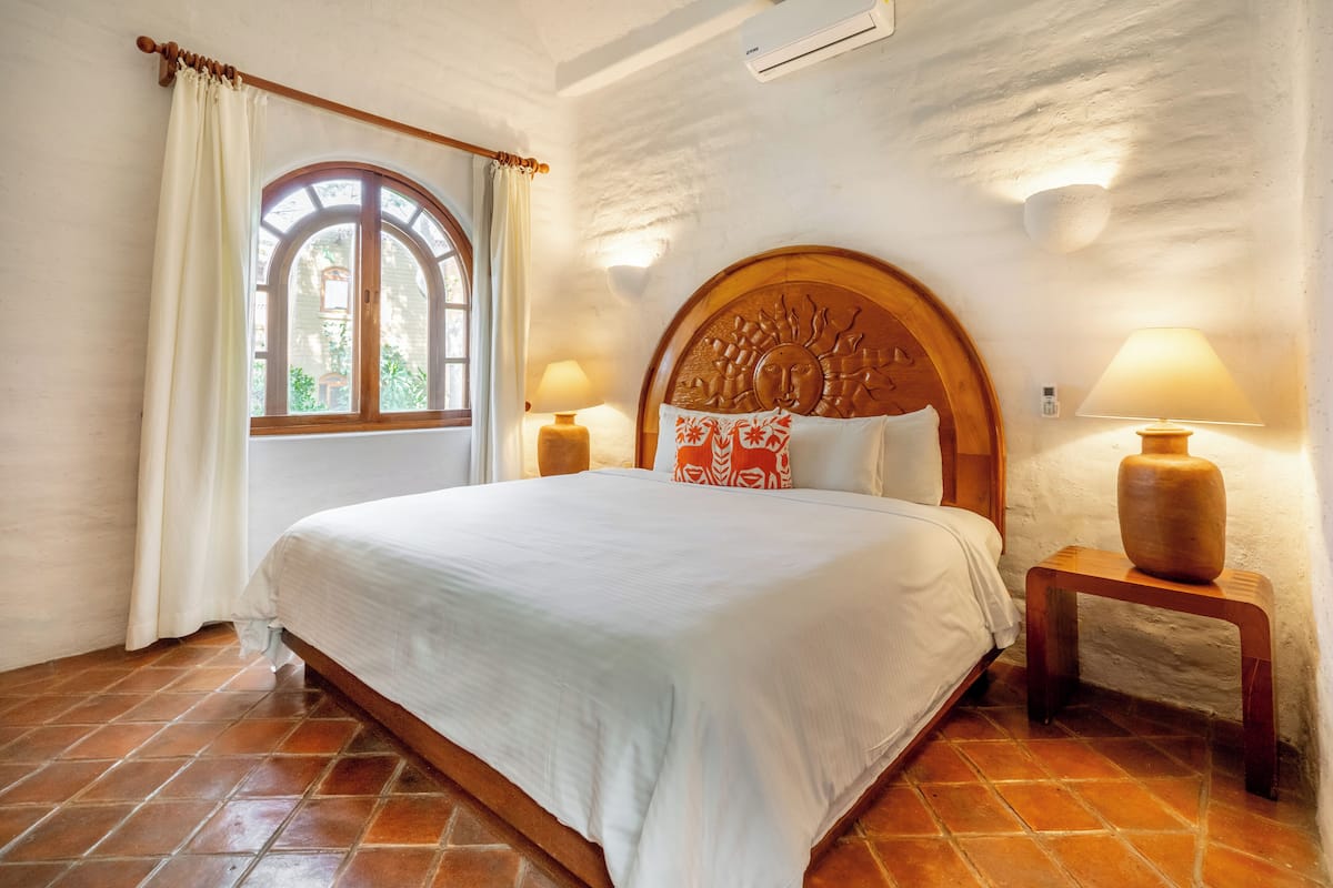 A bedroom with a large bed, two bedside lamps, a window with curtains, and a rustic wall.