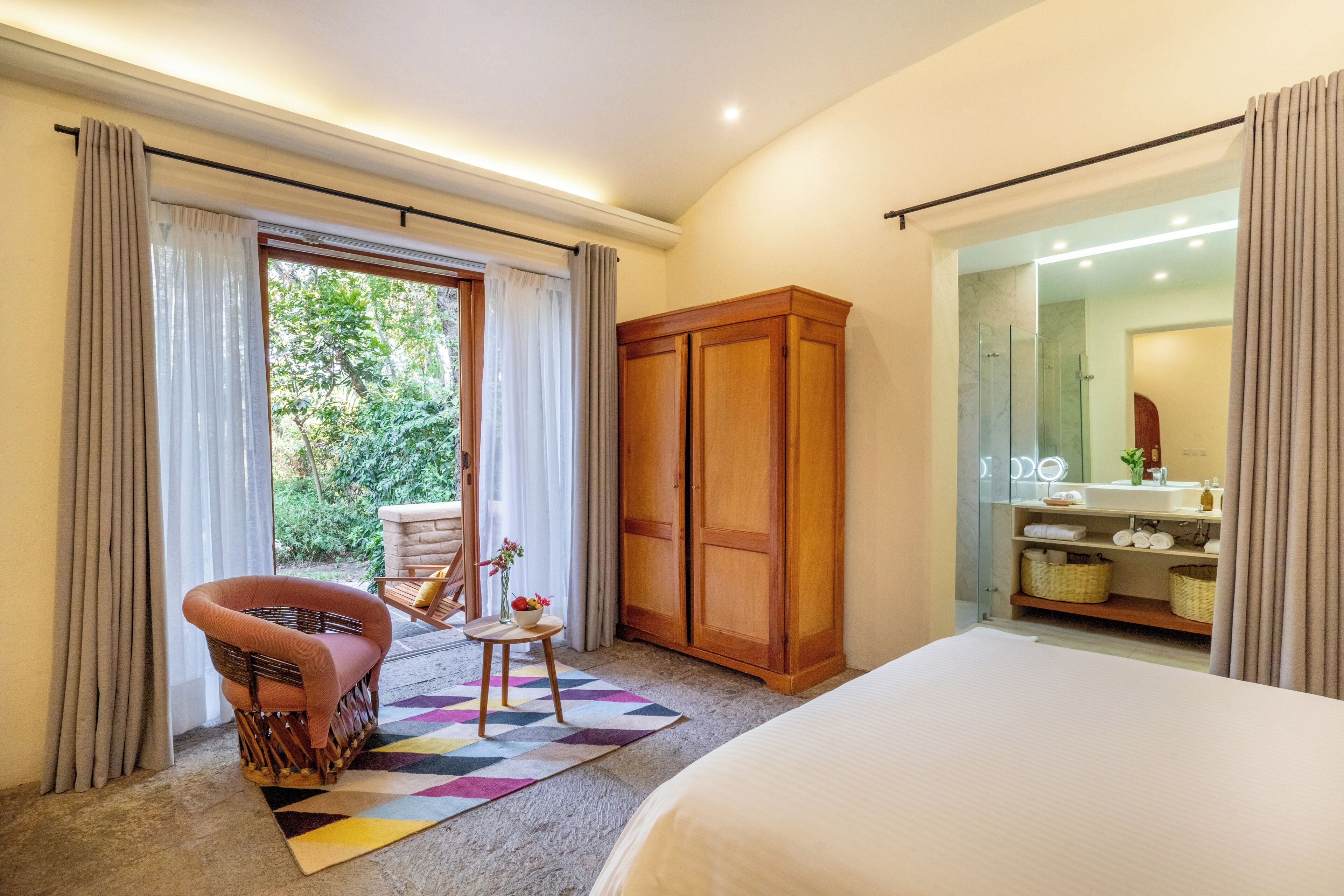 A bedroom with a bed, a wooden wardrobe, a wicker chair, a small table with a vase, and a view of a bathroom.