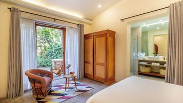 A bedroom with a bed, a wooden wardrobe, a wicker chair, a small table with a vase, and a view of a bathroom.