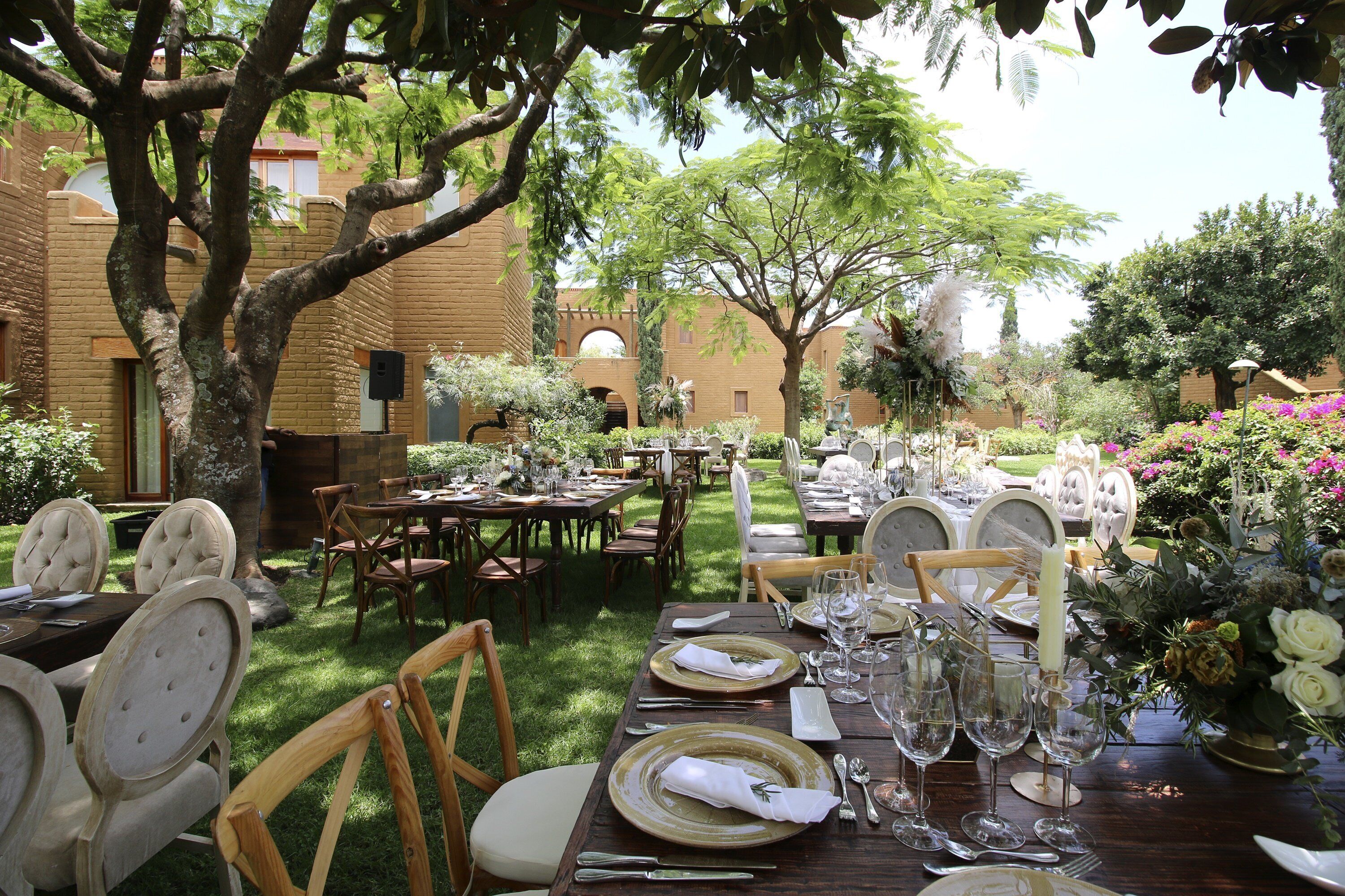 An outdoor dining area with tables set for a meal, surrounded by trees and a brick building.