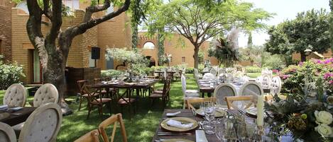 An outdoor dining area with tables set for a meal, surrounded by trees and a brick building.