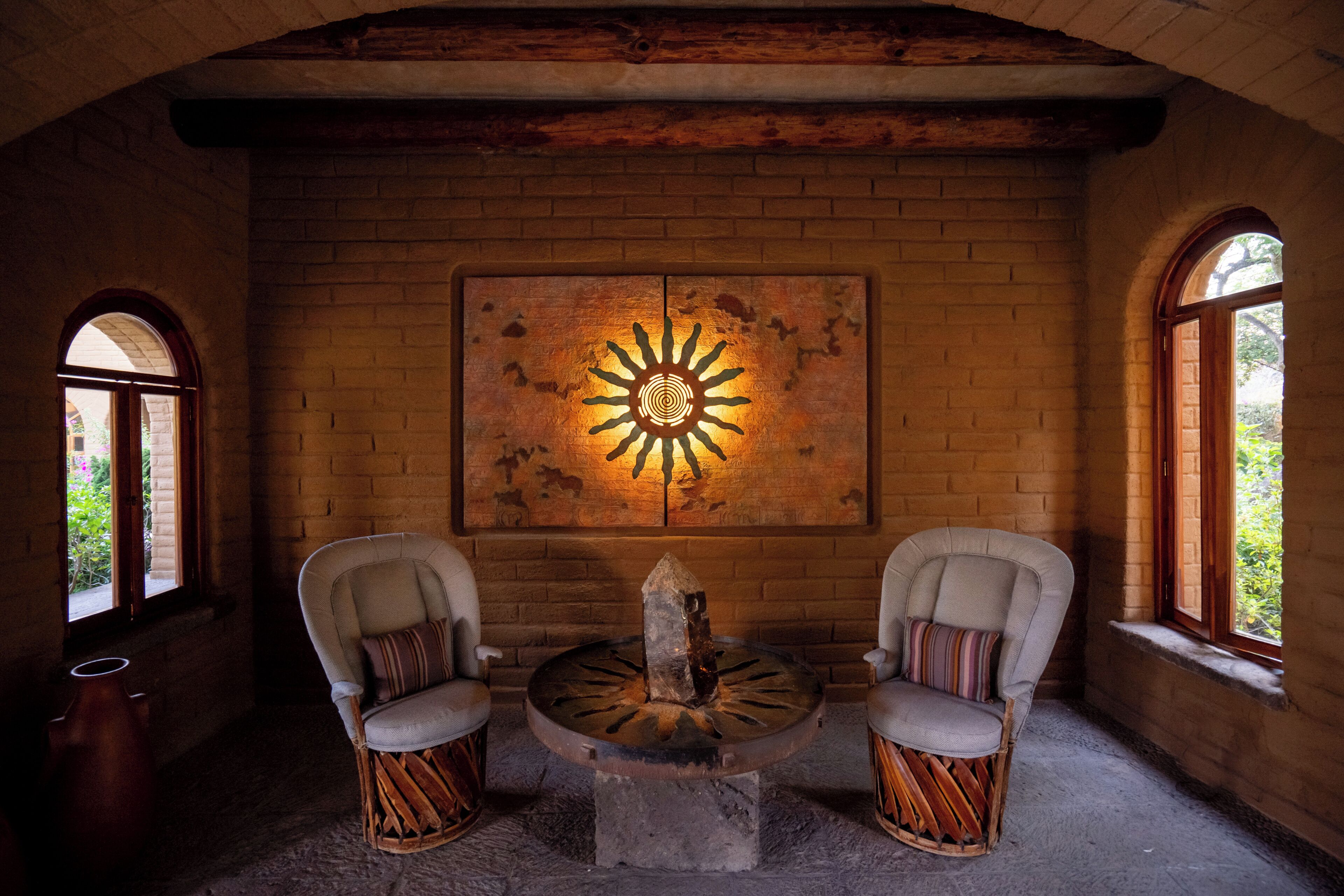 A rustic interior with a brick wall, a sun-like artwork, two upholstered chairs, and a central stone table.