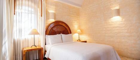 Un dormitorio con cabecera de madera, dos lámparas de noche, una mesita de noche de madera y un ventanal con cortinas.