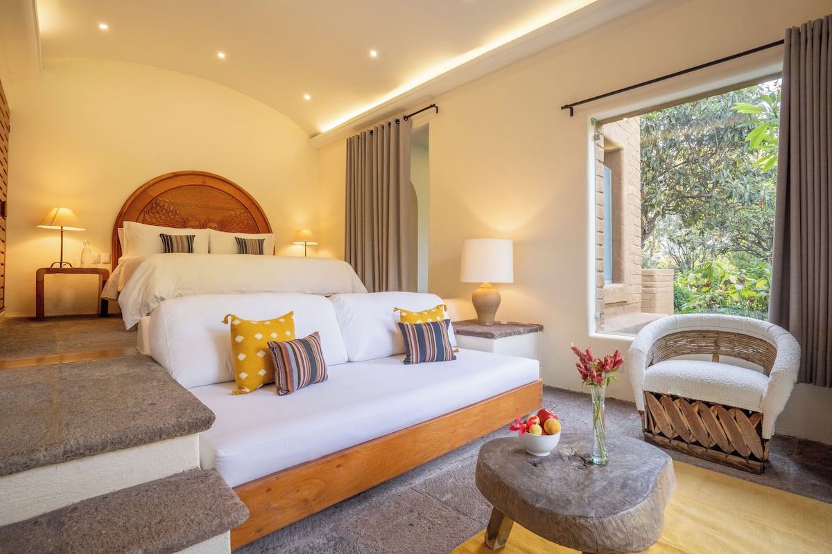 A modern bedroom with a large bed, a wooden headboard, a sitting area with a chair, and a view of greenery outside.
