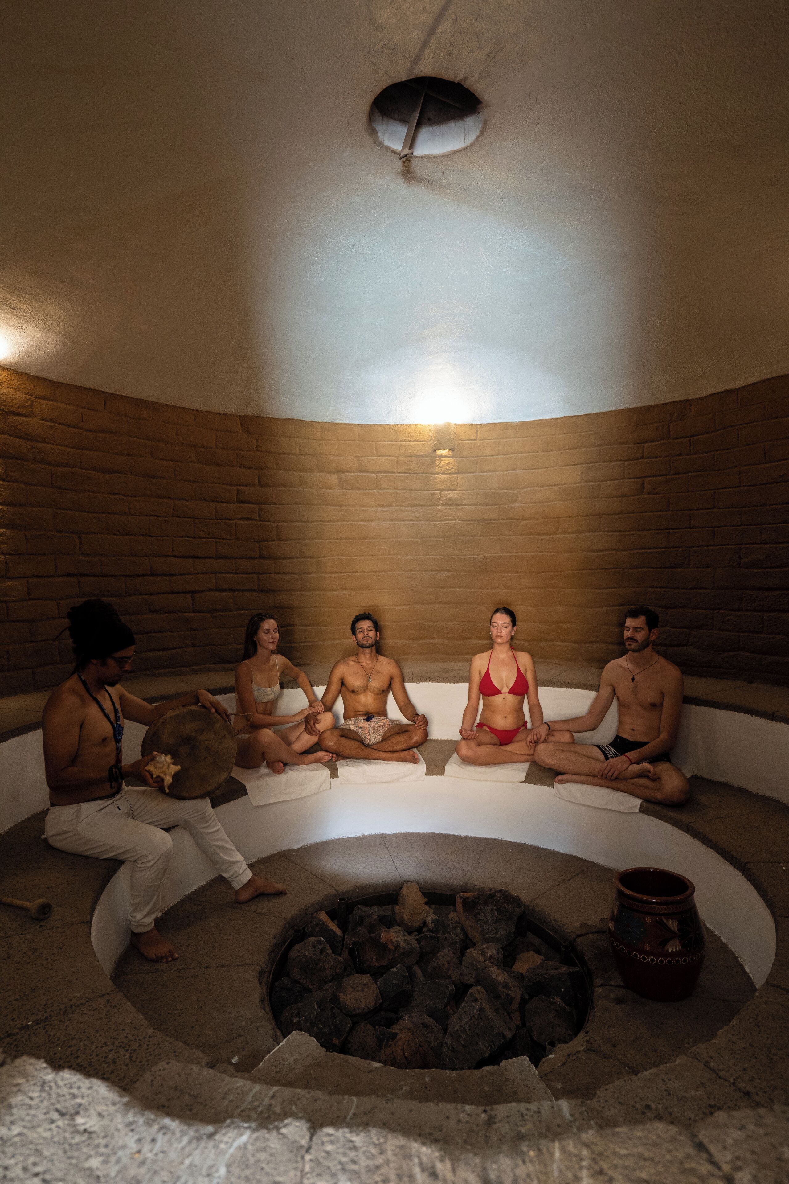 A group of people sitting in a circular arrangement around a fire pit in a room with brick walls and a sloped ceiling.