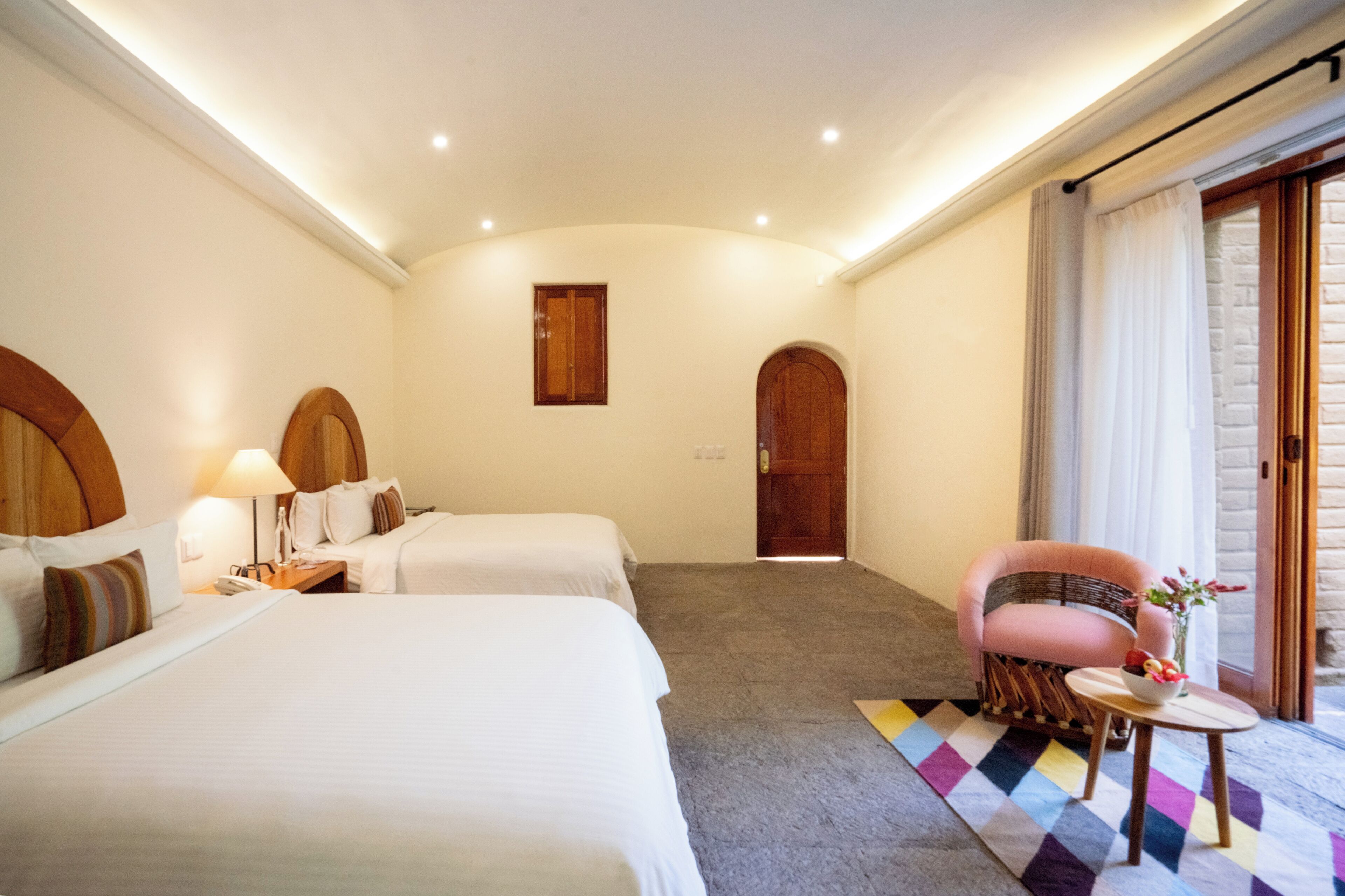 Habitación de hotel con dos camas, una silla, una mesita con un florero y vista al exterior.