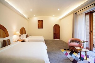 Habitación de hotel con dos camas, una silla, una mesita con un florero y vista al exterior.