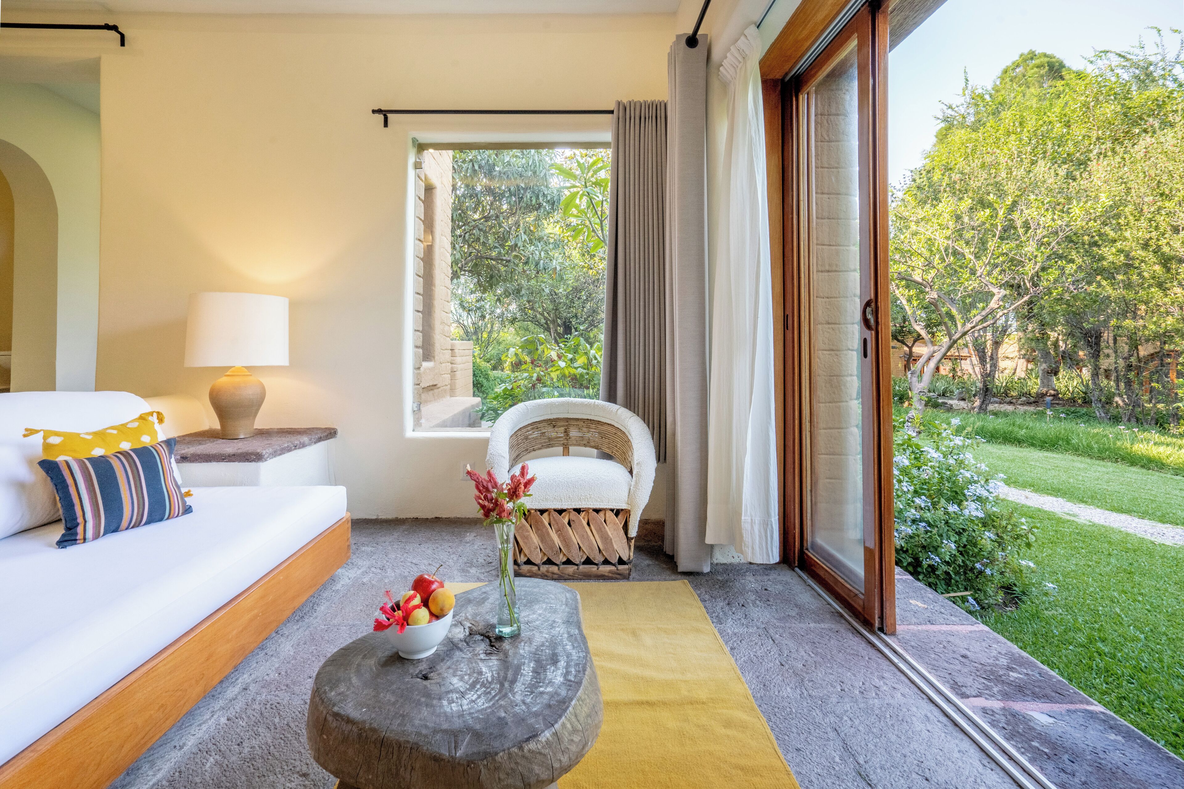 A room with a sofa, a small table with fruit, a chair, and a view of a garden.