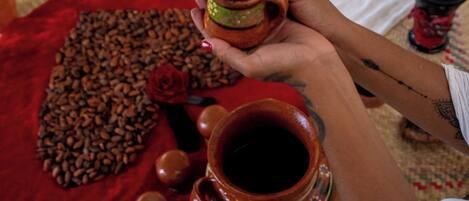 A person holding a small clay pot, with a large clay pot and a red cloth with coffee beans and a rose on the floor.