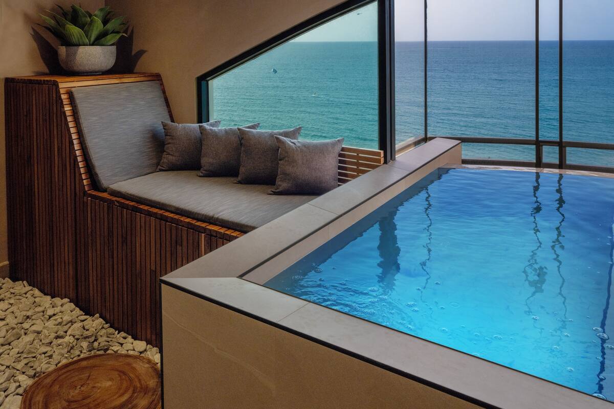 Une piscine à débordement moderne offrant une vue dégagée sur l’océan, un espace détente avec des coussins et un mur d’accent en bois.